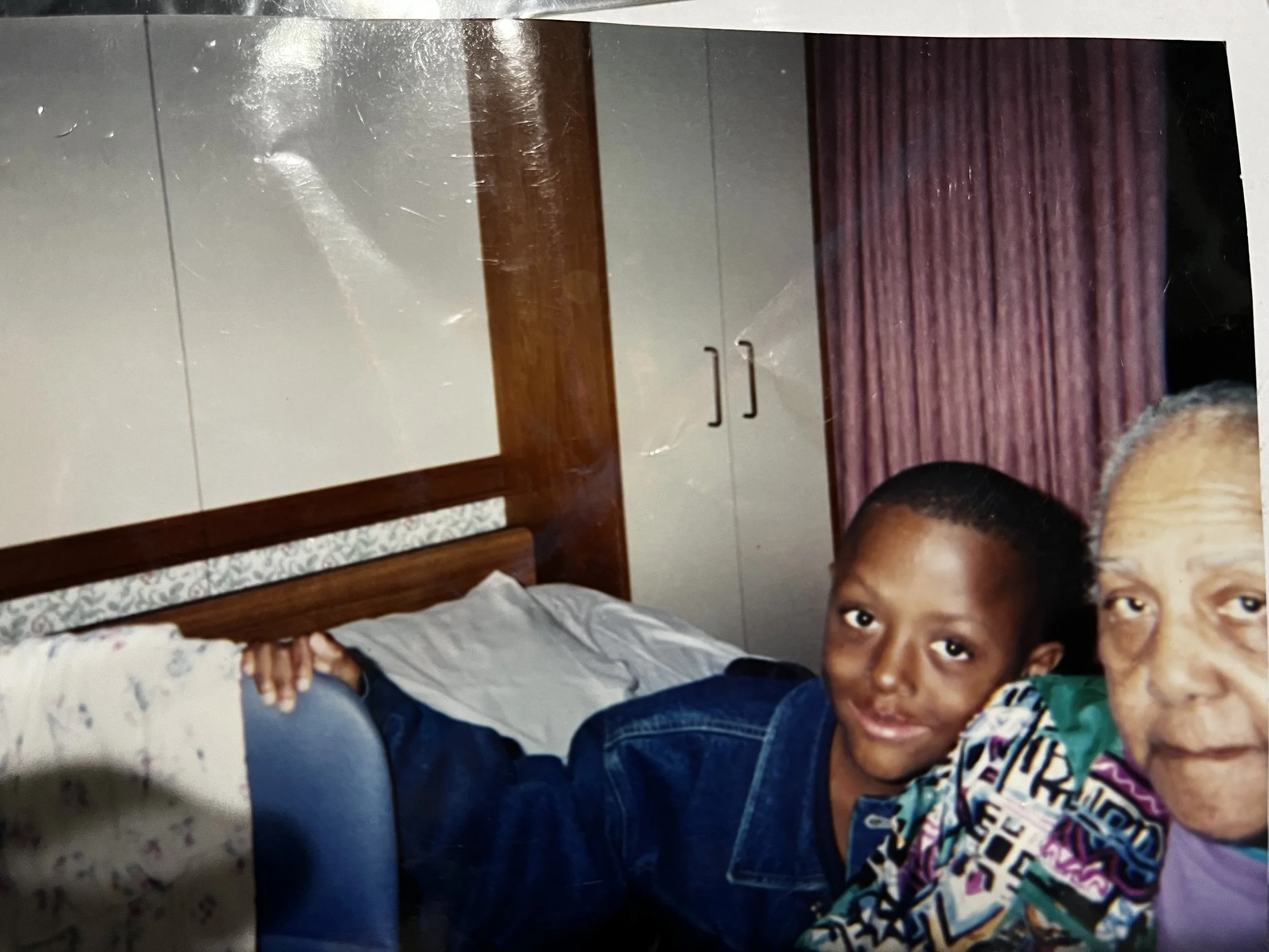 A young boy and an elderly woman are sitting together in a room with a bed, wardrobe, and pink curtains. The boy is smiling and looking at the camera, while the woman is partially visible.