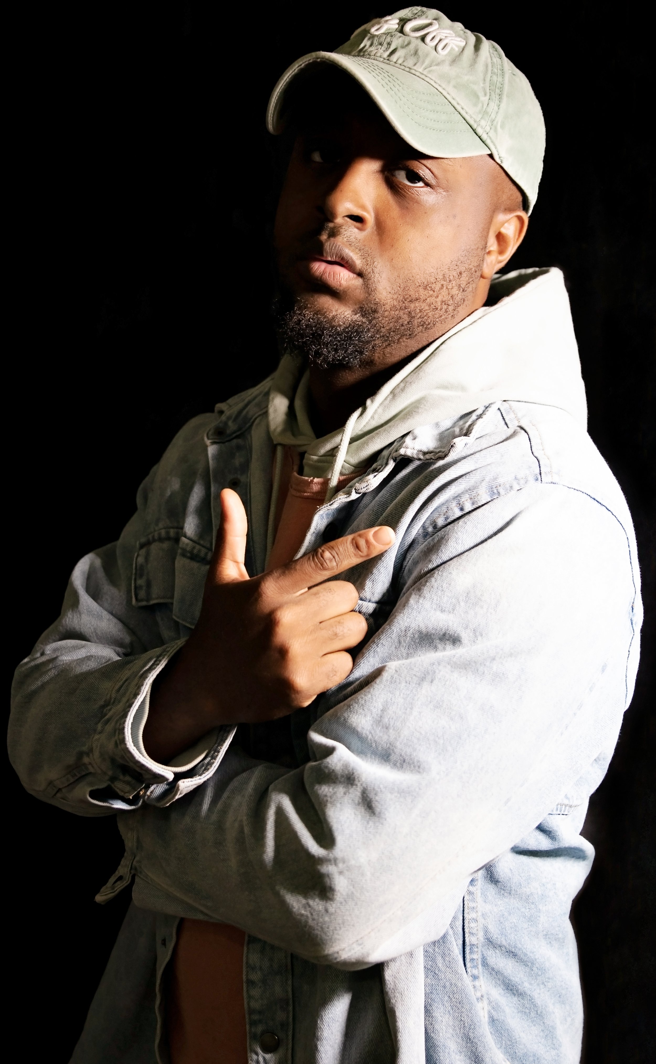 A man wearing a light-colored baseball cap, hoodie, and denim jacket, pointing his finger at his temple while looking into the camera against a black background.