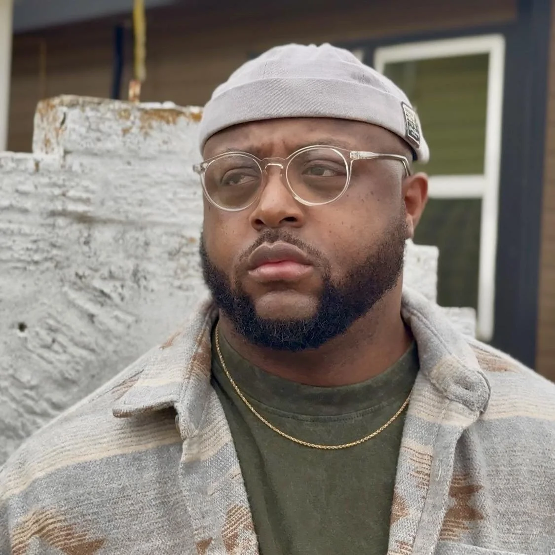 A man wearing glasses, a gray cap, and a beige plaid shirt stands outdoors in front of a weathered white wall and a building with large windows.