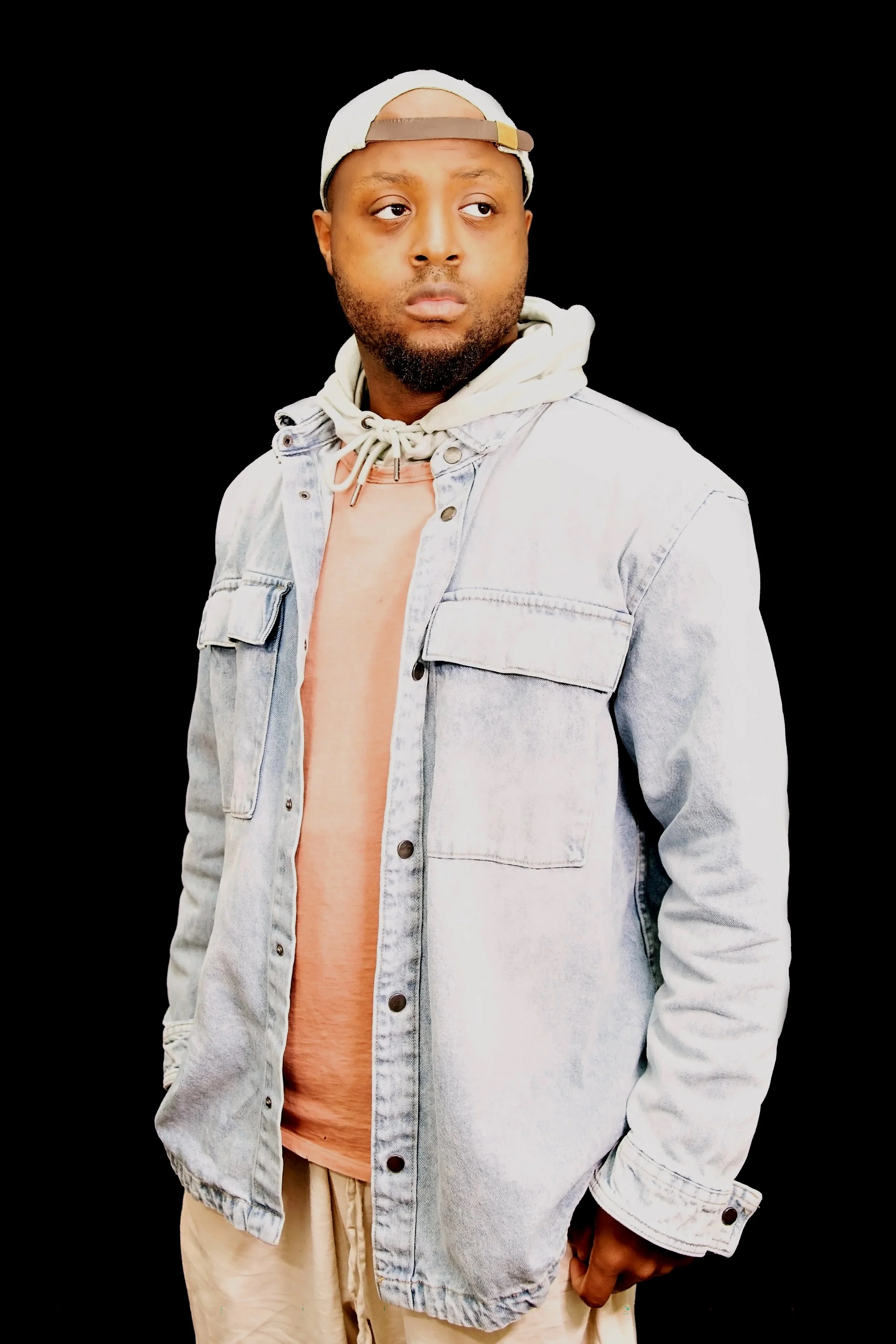 A man wearing a backwards baseball cap, light denim jacket, beige hoodie, and beige pants, standing against a plain black background, looking to the side.