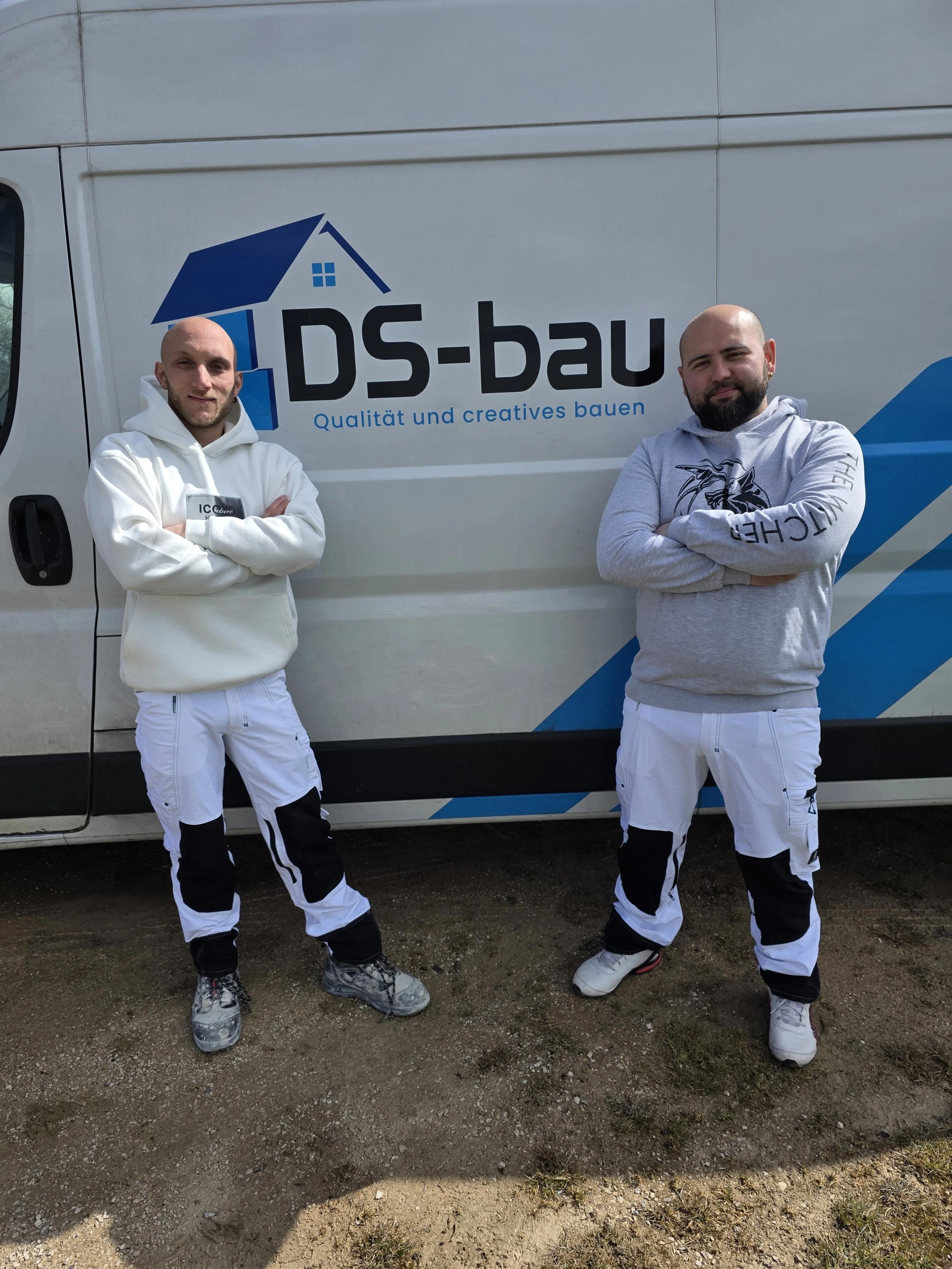 Zwei Männer stehen vor einem weißen Firmenfahrzeug mit dem Logo von DS-bau, beide tragen weiß-schwarze Arbeitshosen und Hoodies, einer in einem weißen Hoodie und der andere in einem grauen Hoodie.