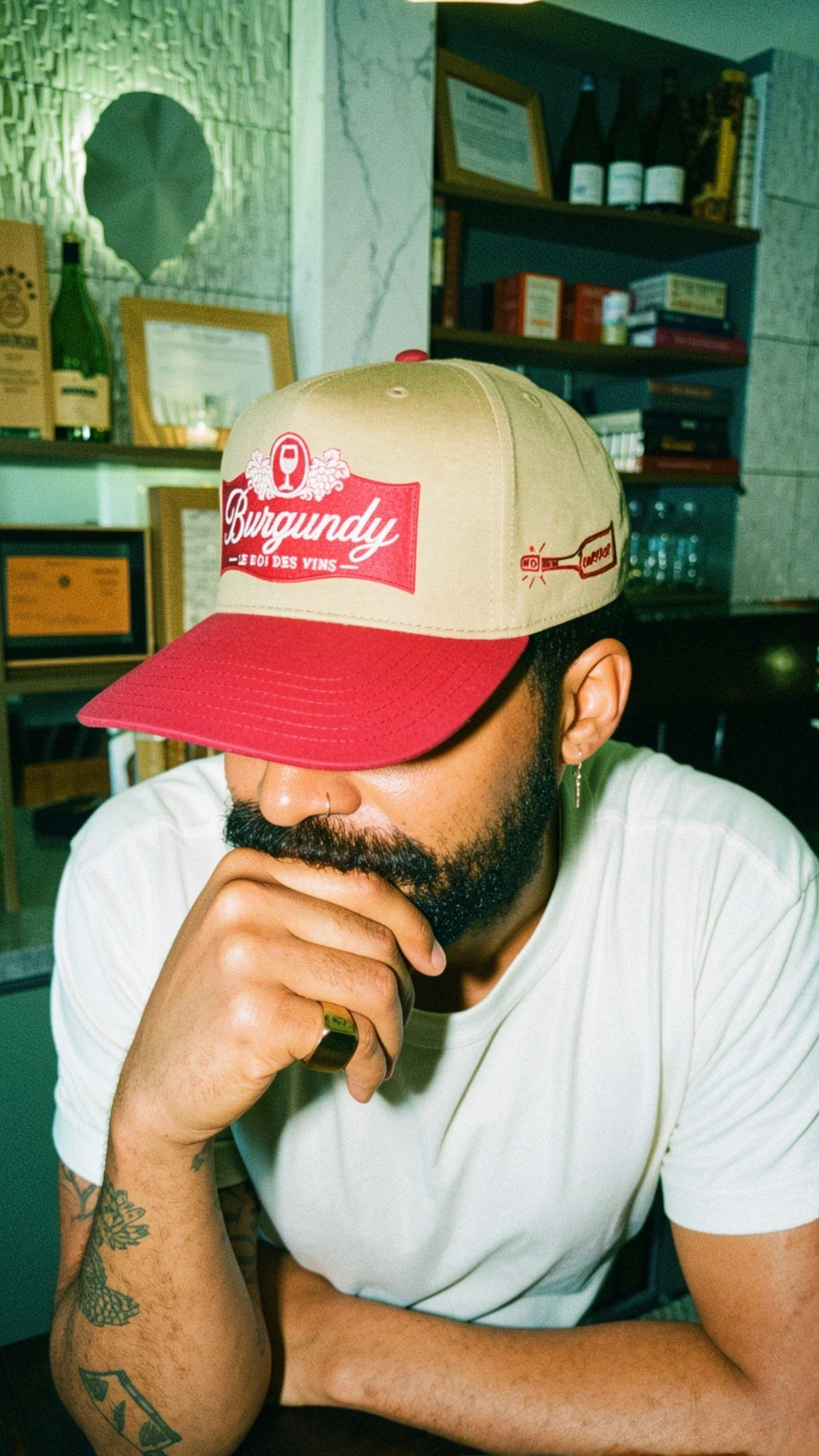 Person with a beard wearing a white t-shirt, a ring, and earrings, has tattoos on arm, resting chin on hand, wearing a cap with red and beige colors, and the brand 'Burgundy' visible on it, in a room with shelves of books and framed certificates.