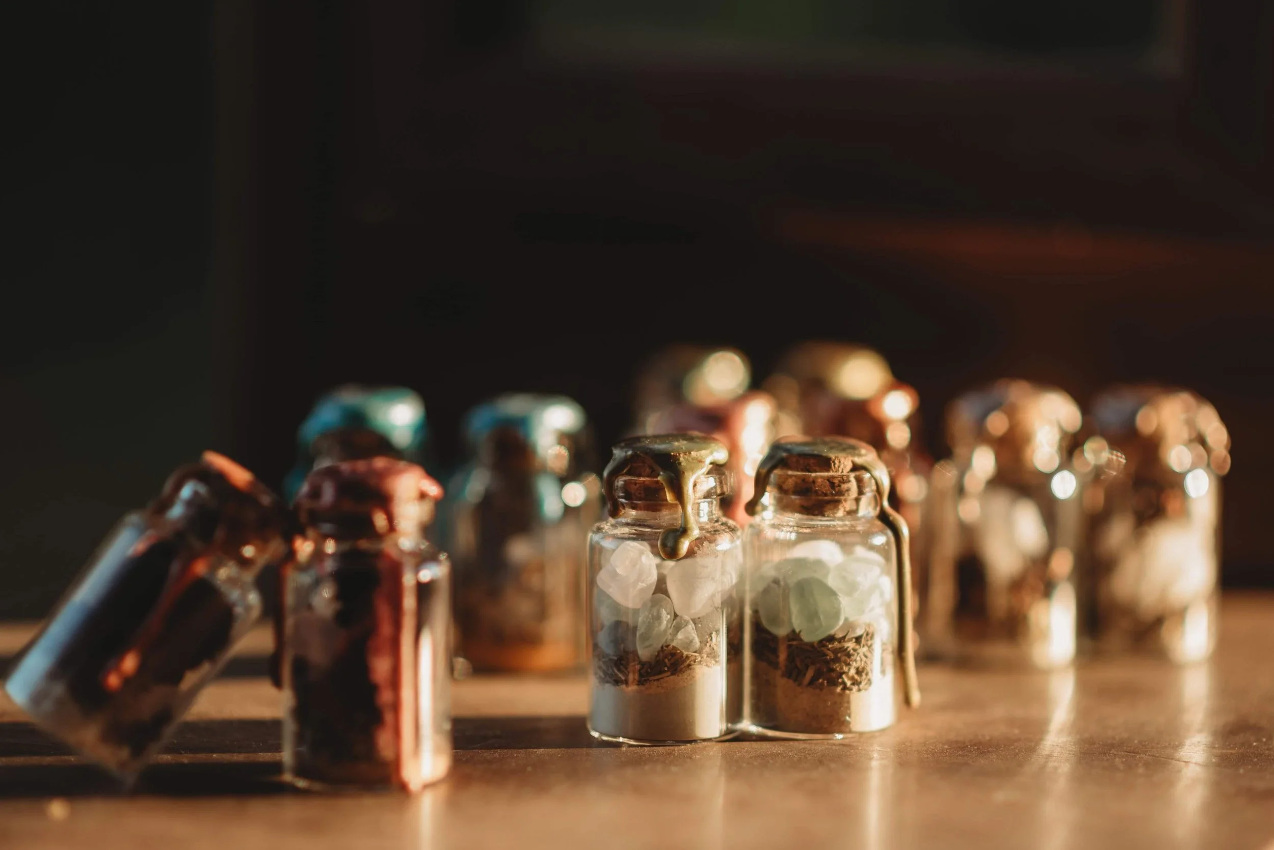 Multiple small glass bottles with wax seal, some filled with dried herbs and others with small stones or crystals, arranged on a wooden surface, with a dark background and warm lighting.