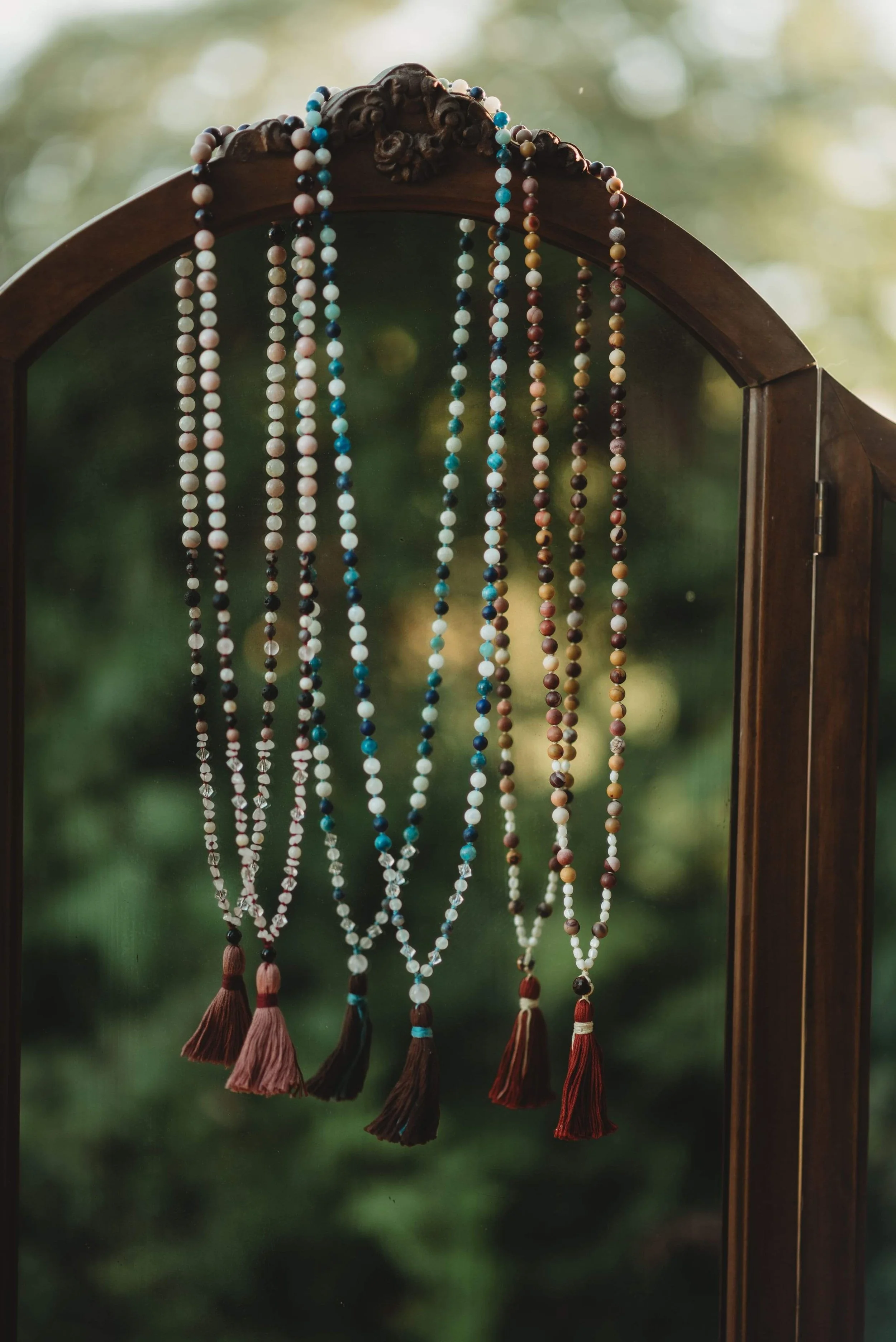 Various beaded necklaces with tassels hanging on a wooden mirror frame outdoors.
