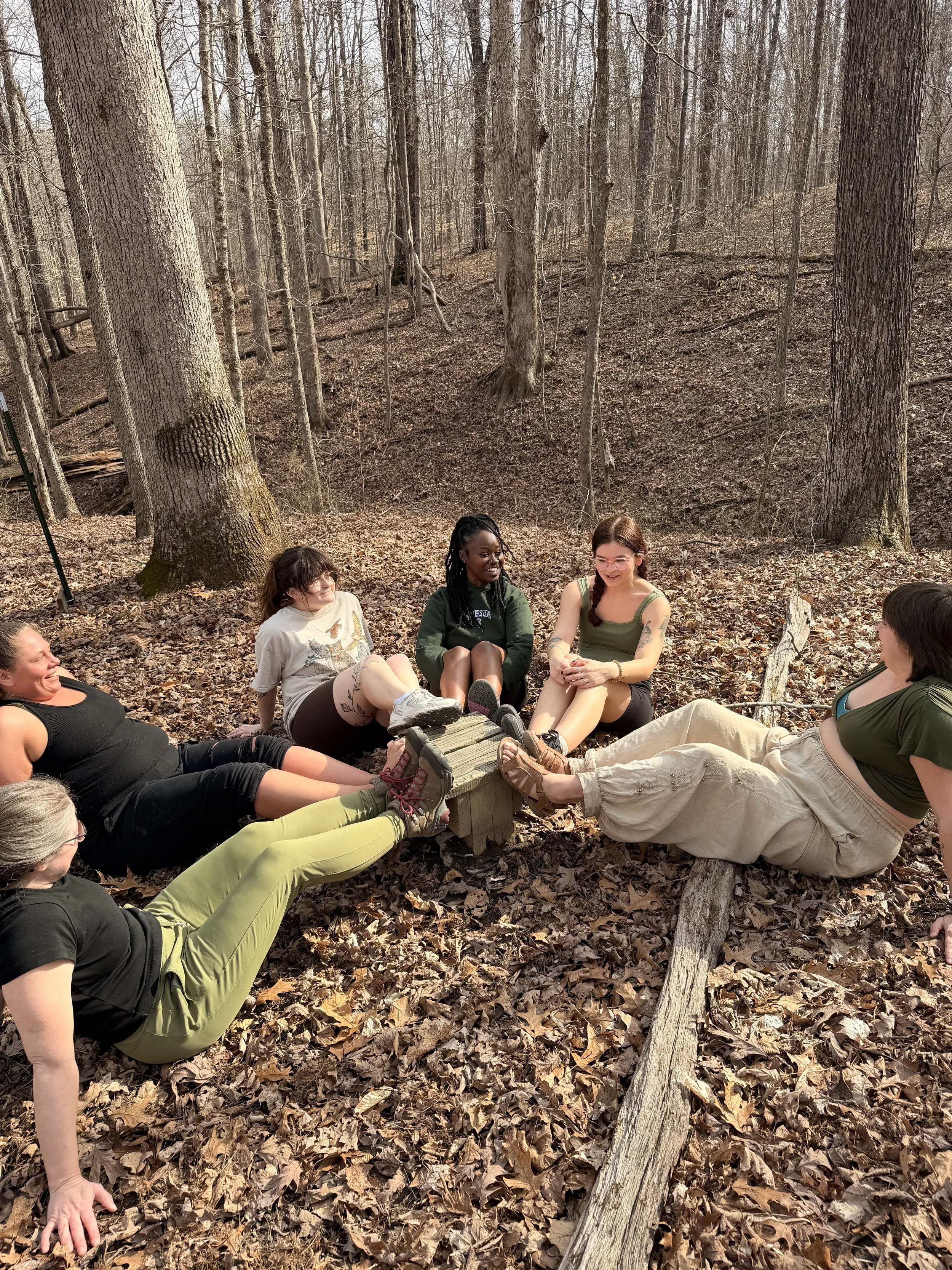 Group of seven young women sitting in circle on fallen leaves in a wooded forest, with their feet placed on a small wooden platform as a part of the low challenge course at CYO Camp Rancho Framasa.
