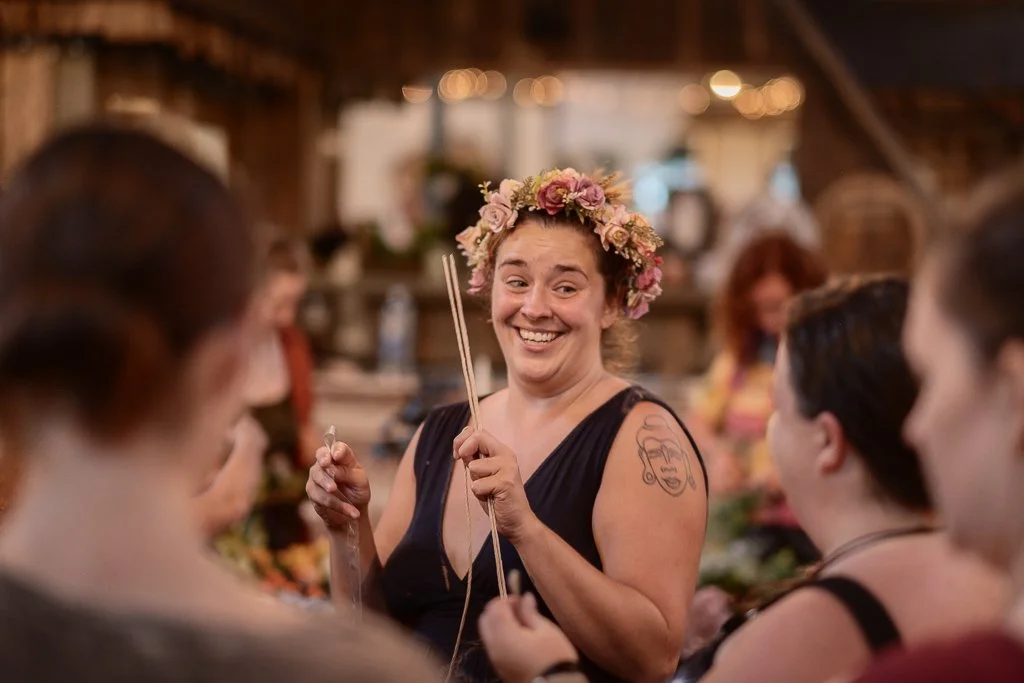 Woman wearing a flower crown smiling at a gathering of people in a rustic indoor setting.