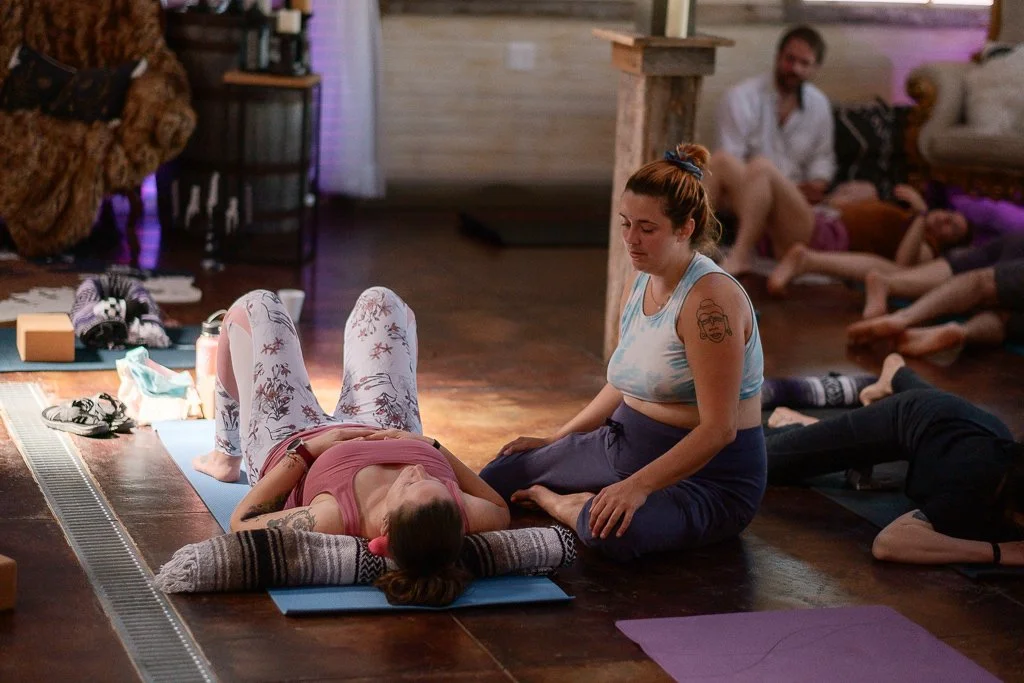A woman lying on a yoga mat in a restorative yoga pose with her eyes closed, while an instructor sits beside her in a cross-legged position, in a cozy indoor setting with other people resting on mats in the background.