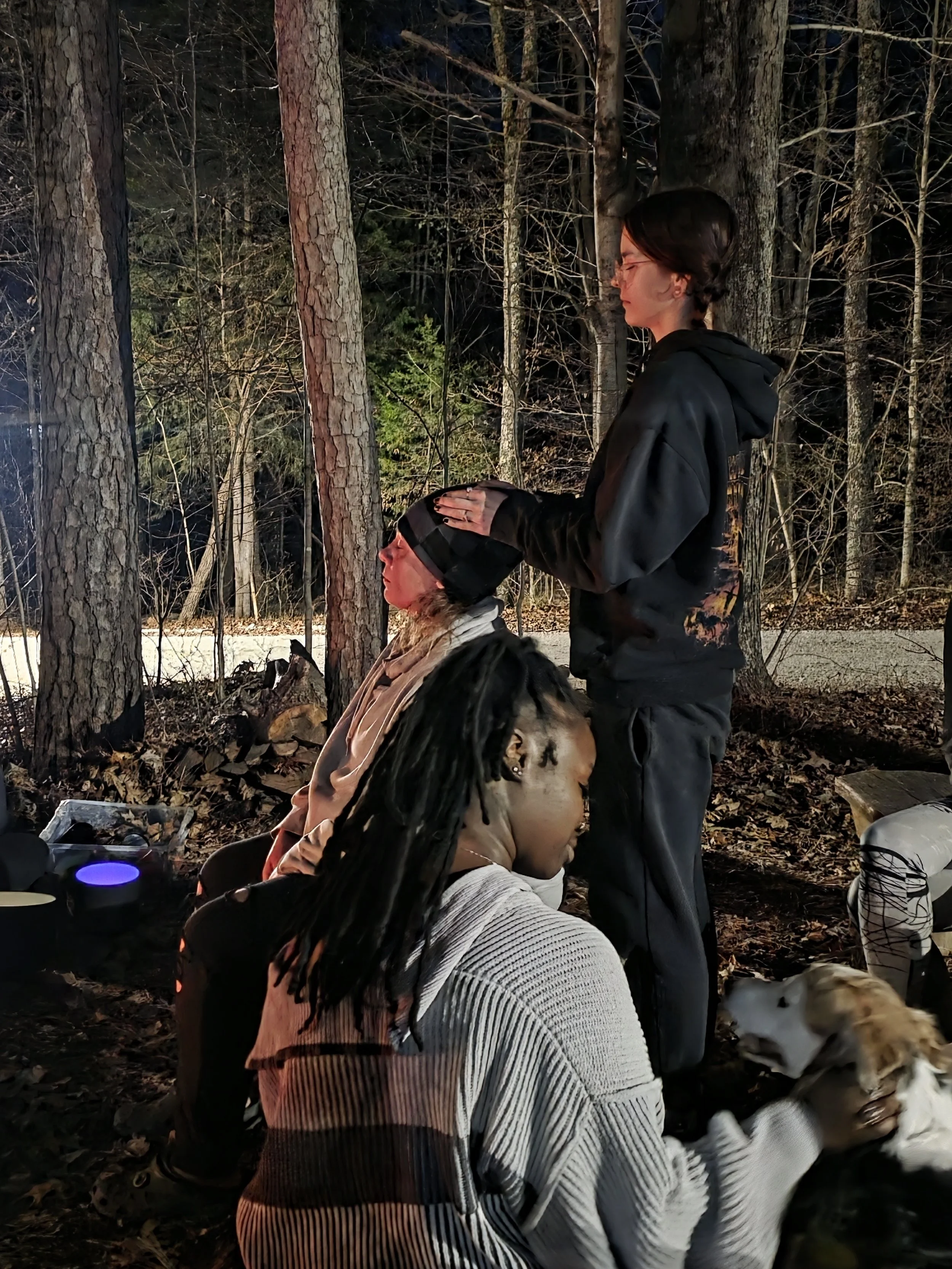 Group of people gathered outdoors in a wooded area at night, with one person standing and gently touching the forehead of another person sitting with eyes closed, while a person with dreadlocks sitting nearby holds a dog. The scene is illuminated by outdoor lighting.