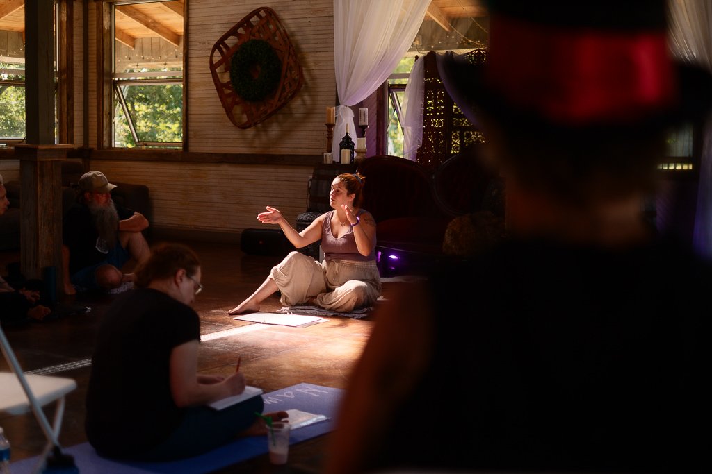 A woman sitting on a rug, speaking or performing in front of an audience in a cozy, rustic room with wooden walls and windows, while others sit on the floor listening.