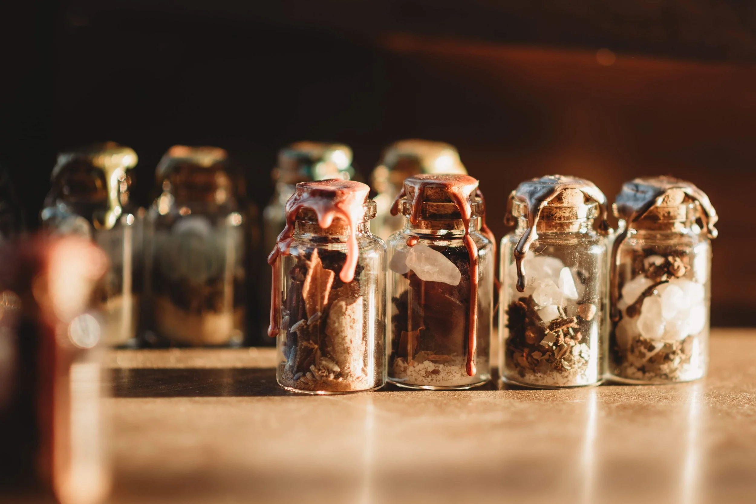 Small glass jars filled with crystals and herbs, with some wax spilling over the edges, illuminated by warm sunlight.