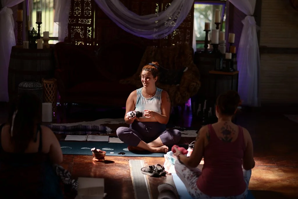 A woman in a light blue tank top and leggings sitting cross-legged on a yoga mat, gesturing with her hands, leading a yoga or meditation class with three women in a dimly lit room with candles and draped fabric.