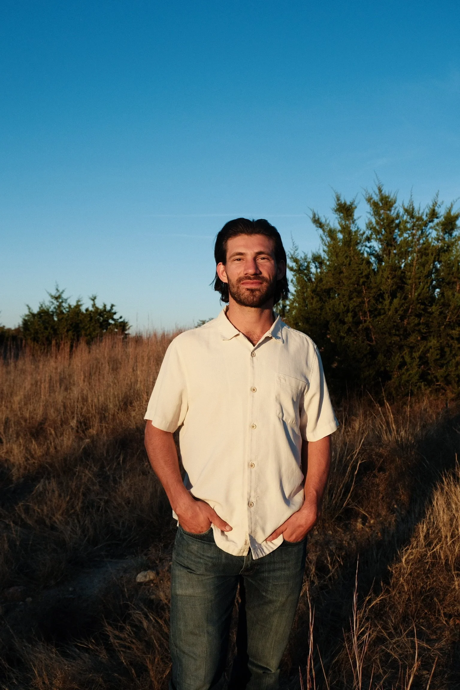 A man standing outdoors in a field during sunset with his hands in his pockets, wearing a light-colored short-sleeve shirt and jeans, with trees and a clear blue sky in the background.