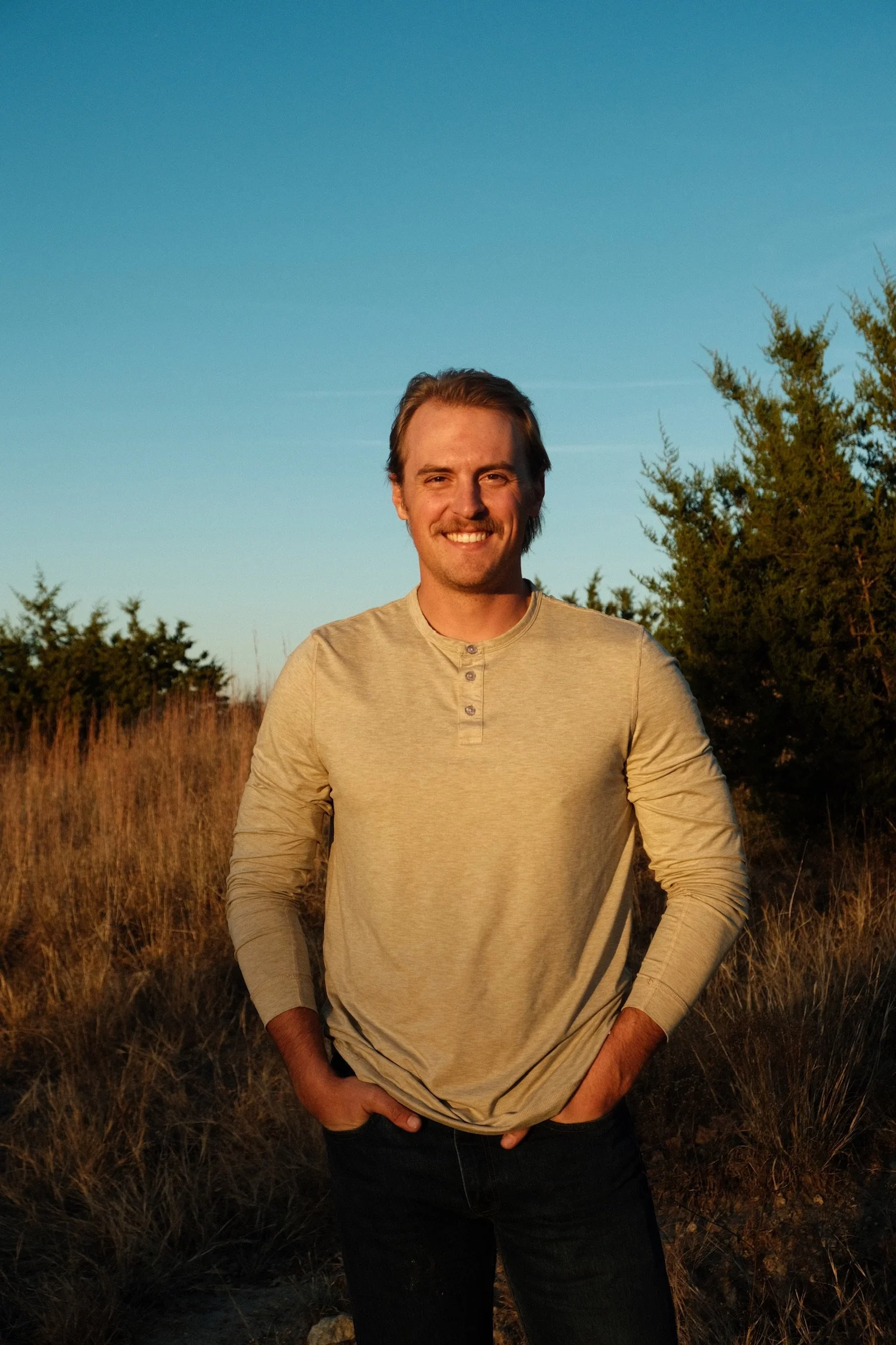A smiling man standing outdoors in a rural area, with dry grass and trees, under a clear blue sky.