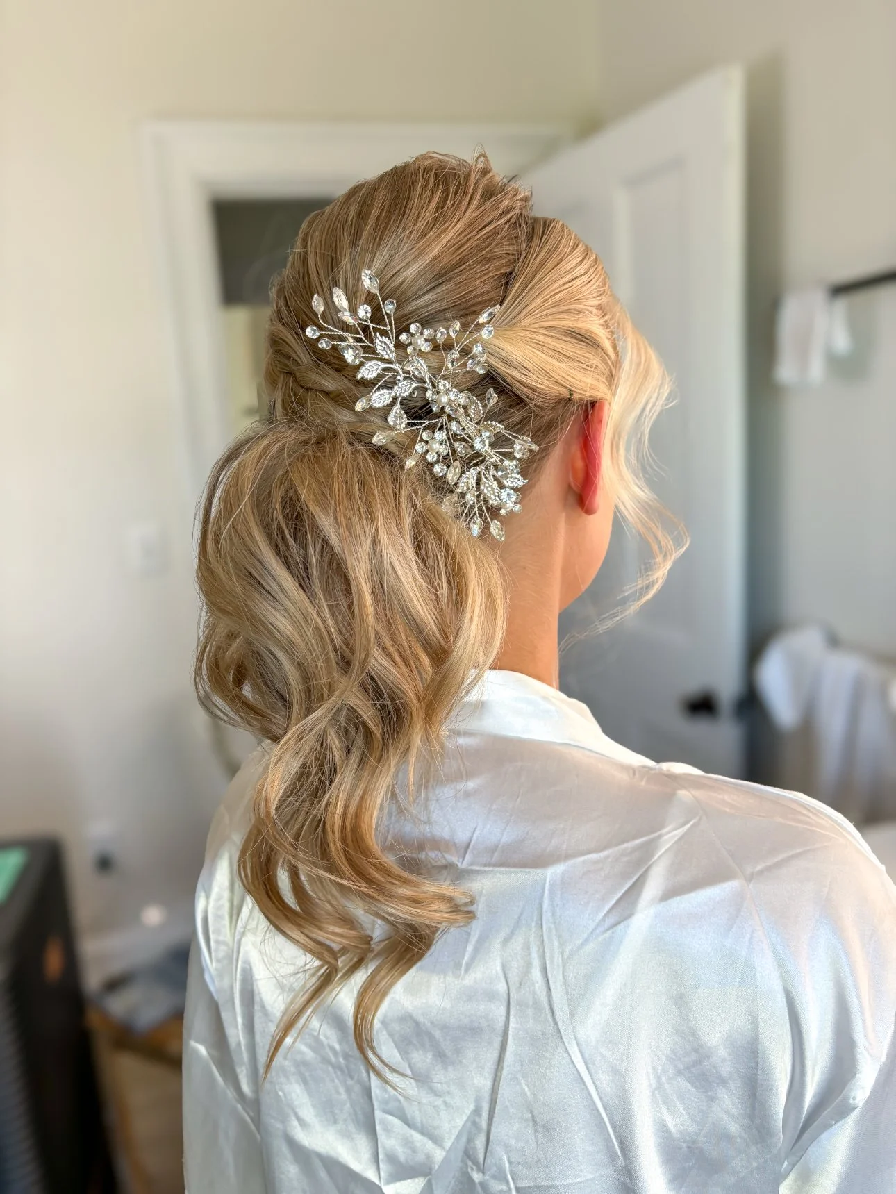 A woman with blonde hair styled in loose waves wearing an ornate silver hair accessory with leaf and floral designs, seen from behind.