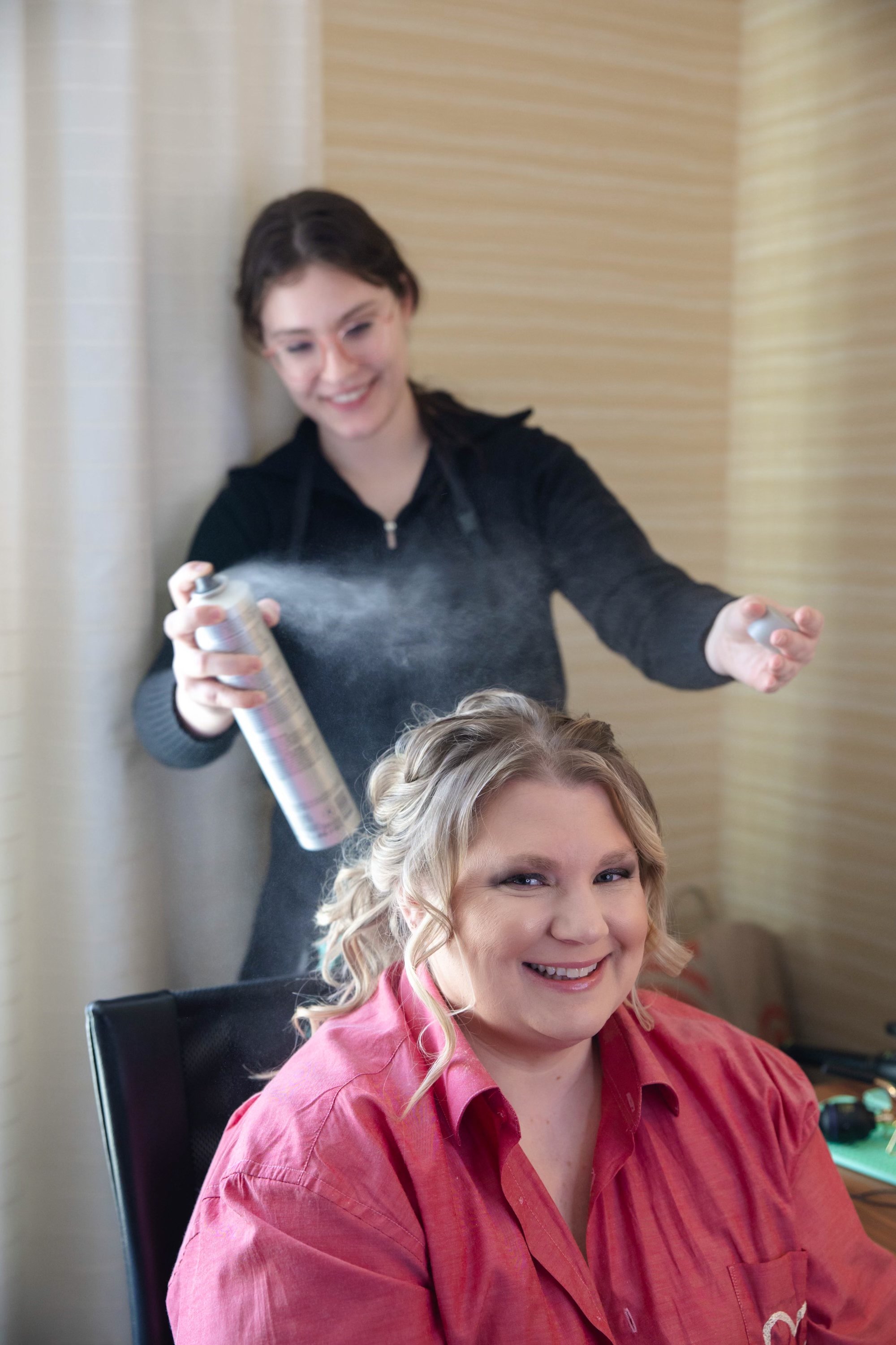 A woman with blonde curly hair smiling at the camera while another woman sprays hair spray on her hair in a salon or home setting.