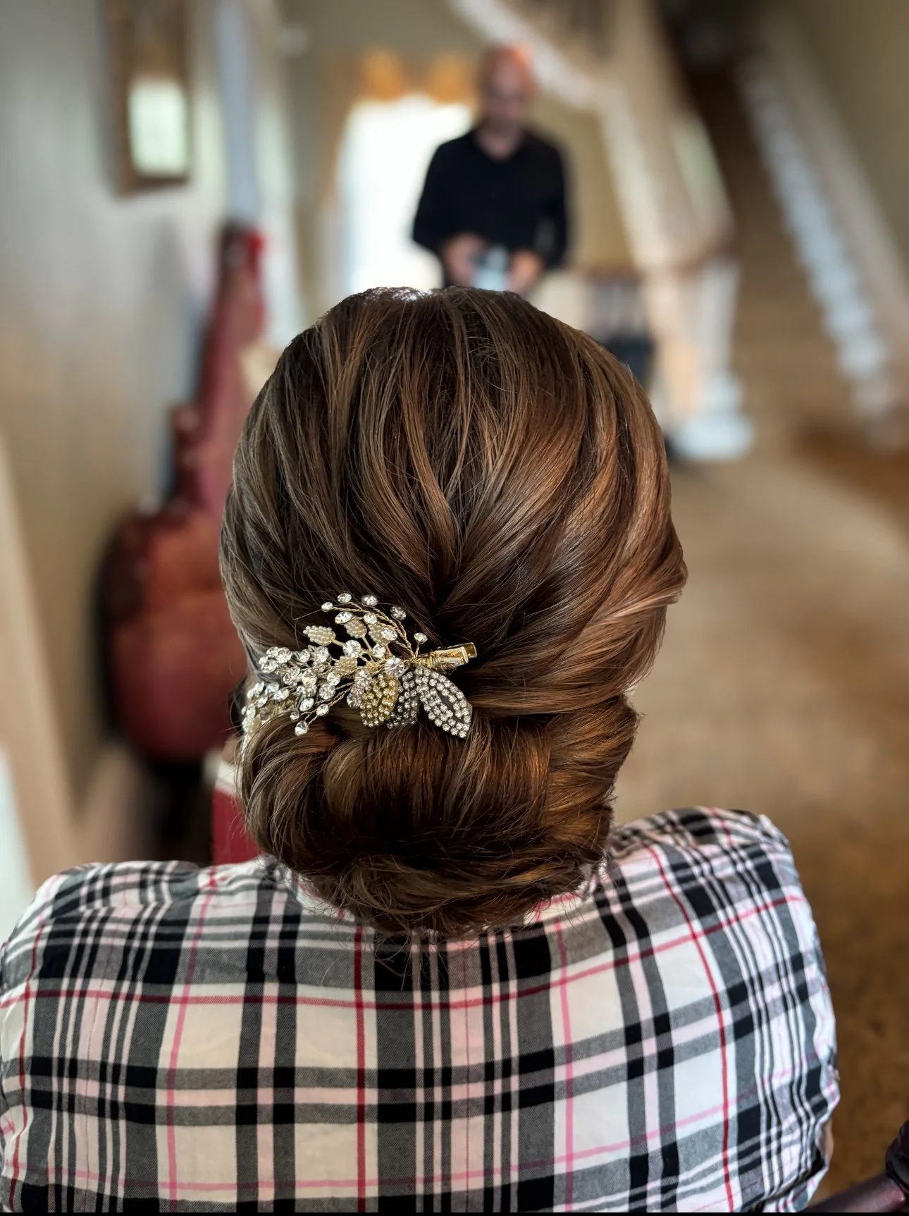 Back of woman's head with styled brown hair and a decorative rhinestone hair clip, wearing a plaid shirt, in an indoor setting with blurred background.
