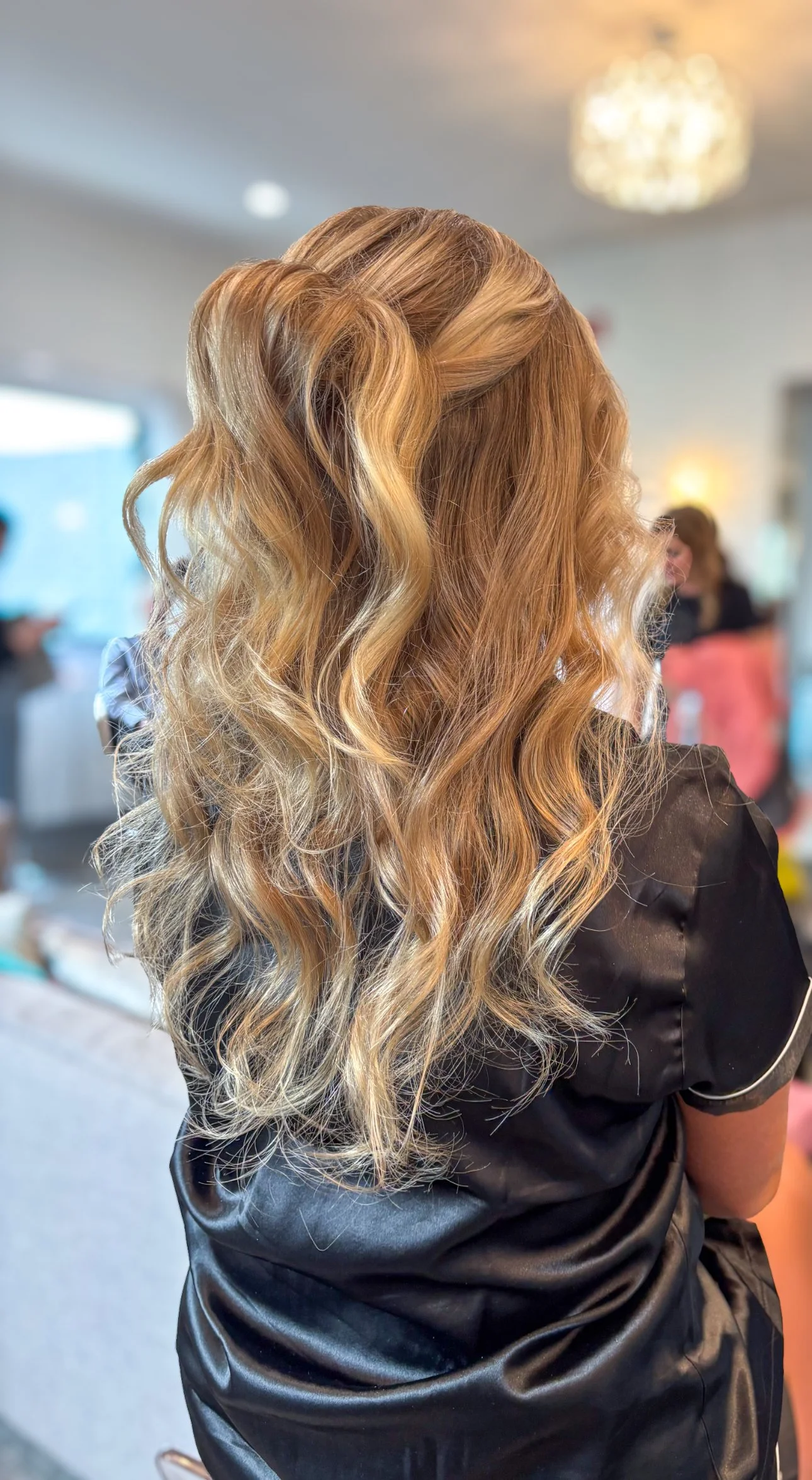 Back view of a woman with long, wavy, blonde hair styled in loose curls, wearing a black satin top, in a brightly lit indoor space with blurred background of people.