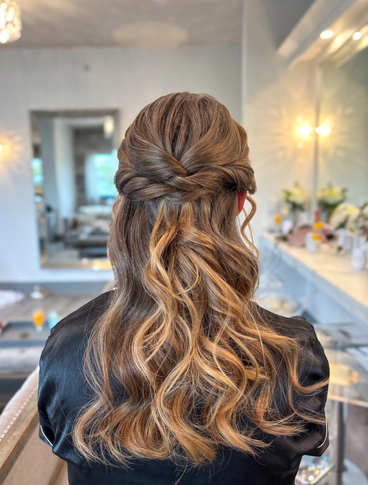 Back of a woman's head with styled, wavy, ombre hair in shades of brown and blonde, in a modern, well-lit room with flowers and decorative items.
