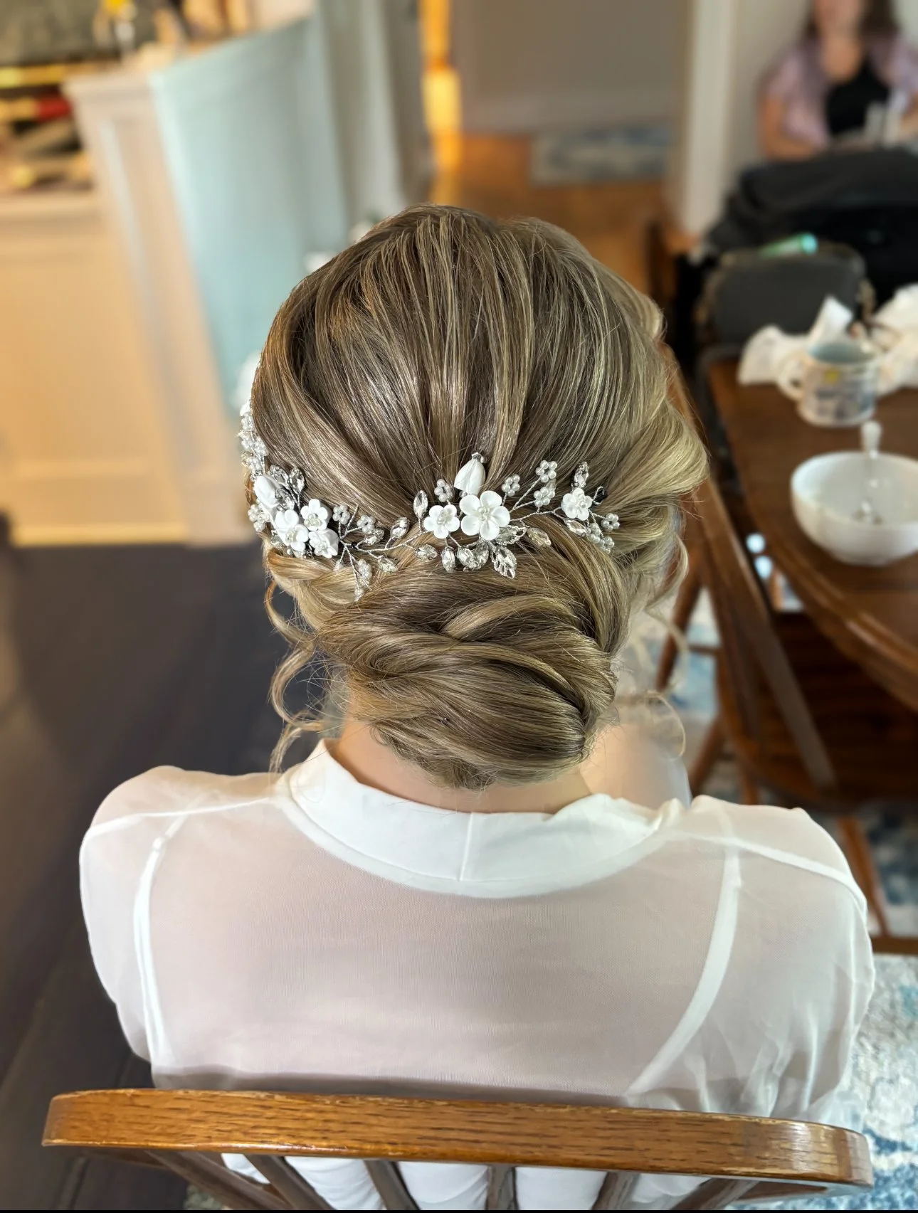 A woman with styled blonde hair wearing a white shirt and an elegant floral hair accessory, sitting on a wooden chair in a home setting.