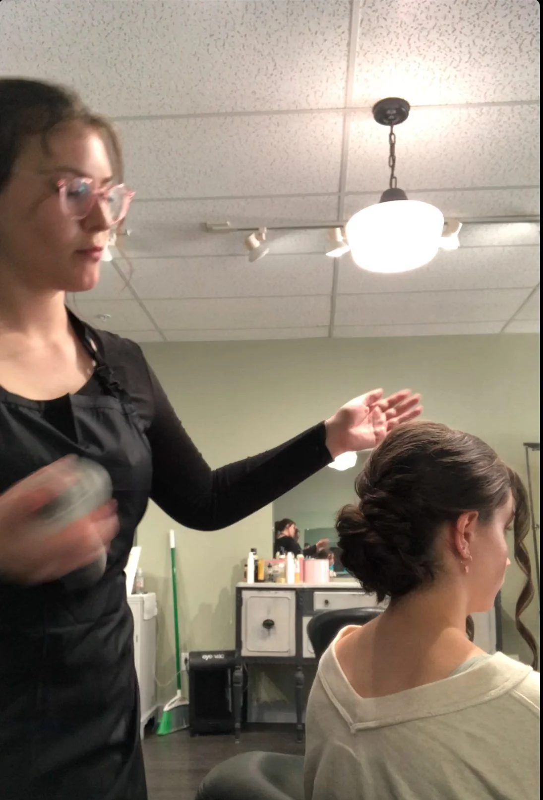 A hairstylist working on a woman's hair in a salon, with salon products on a counter in the background.