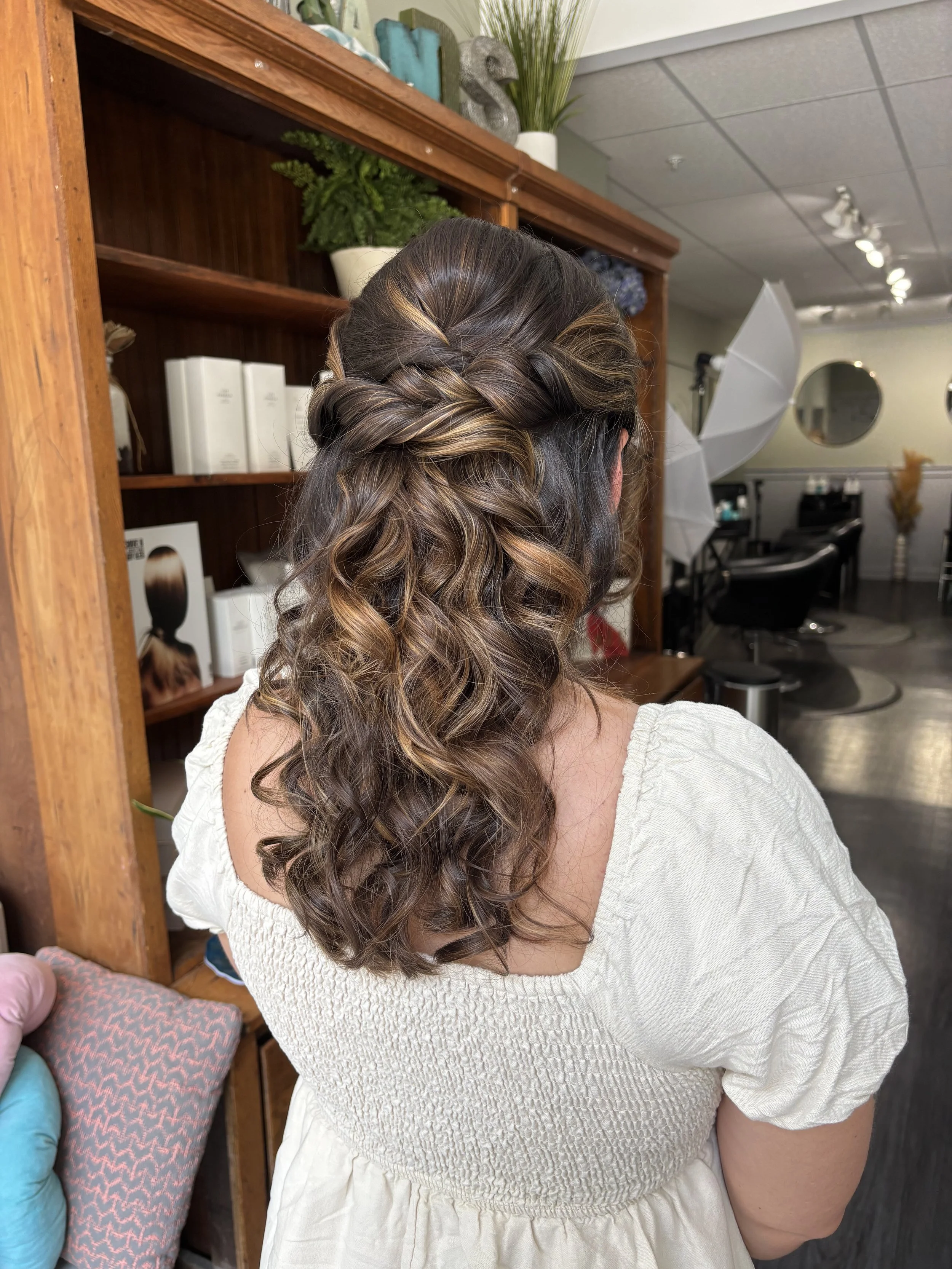 Back view of a woman with shoulder-length curly hair with highlights, wearing a beige top, in a salon with hair products and equipment in the background.