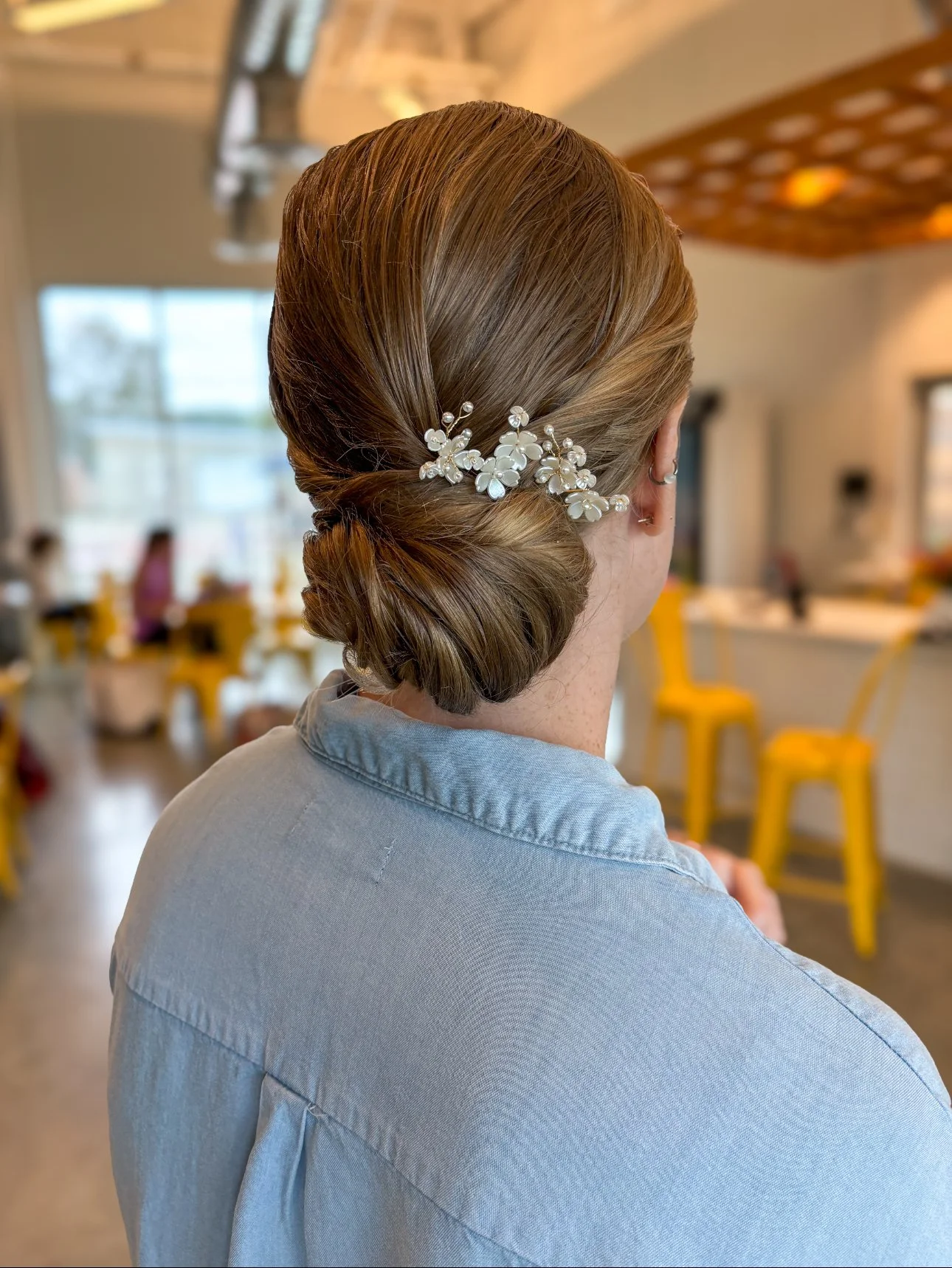 Back view of a woman with styled hair and a decorative hair accessory, wearing a light blue shirt, in a bright indoor setting with yellow chairs and other people in the background.