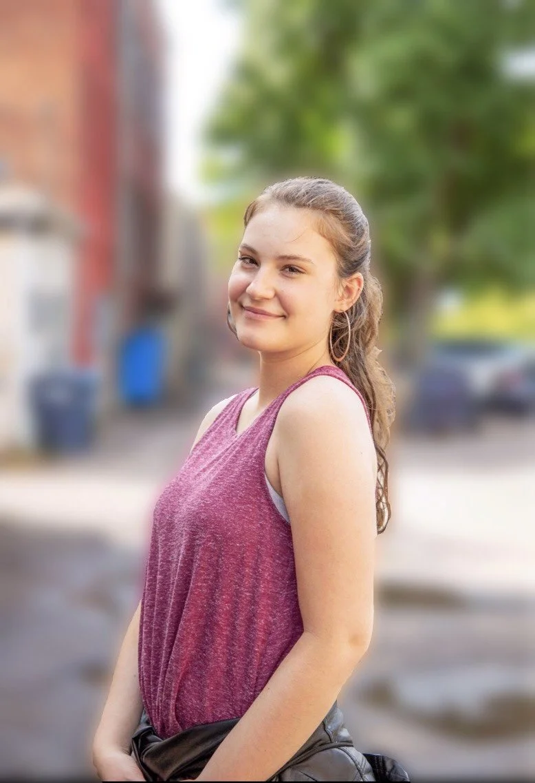 Young woman with light brown hair in a ponytail, wearing a sleeveless pink top, standing outdoors with blurred background of trees and buildings, smiling at the camera.