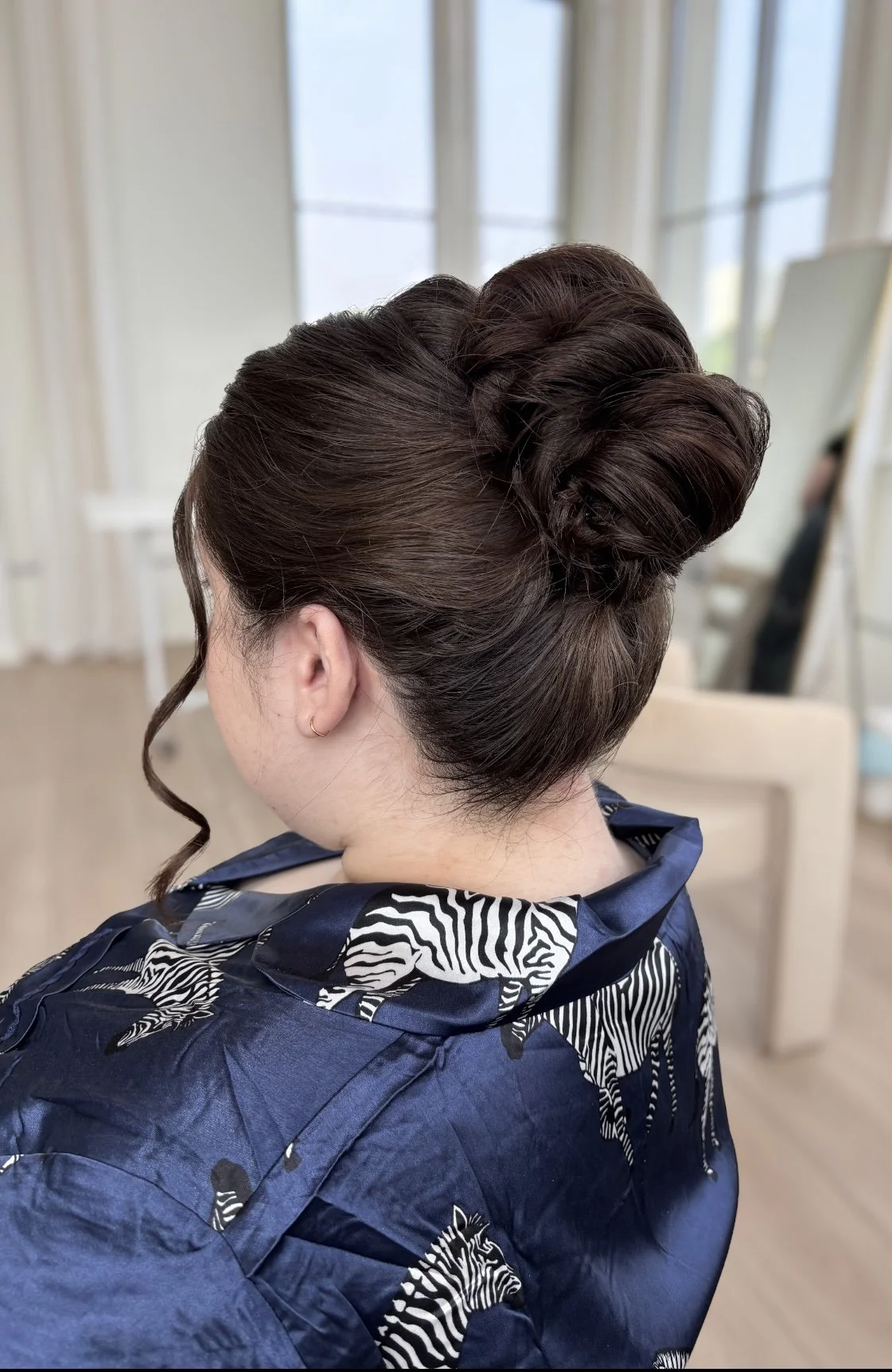 Back of a woman's head showing an elegant updo hairstyle, with a curl hanging down on the side, in a room with large windows and a mirror in the background, wearing a navy blue garment with zebra print.