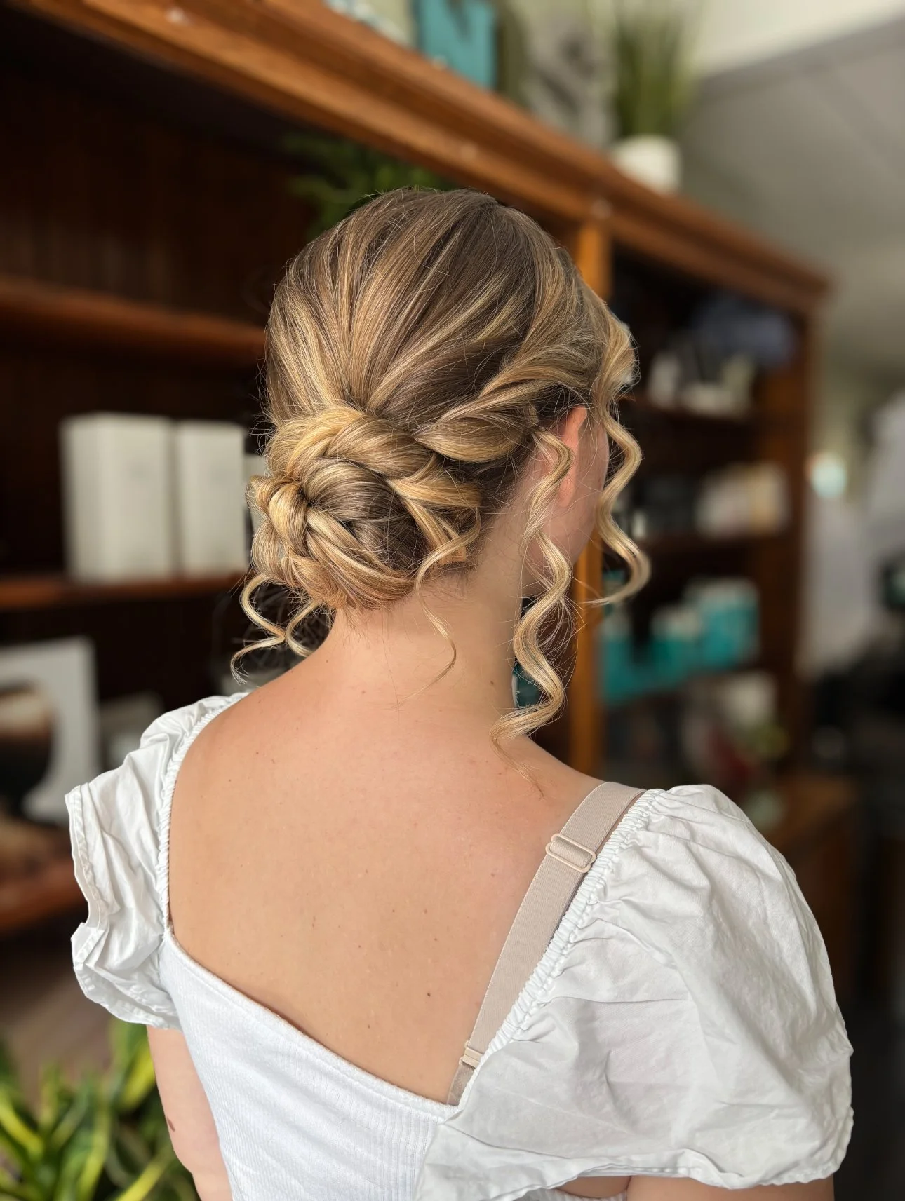 Back view of a woman with styled blonde hair in an intricate updo, wearing a white top with puffed sleeves and beige bra straps, standing indoors with wooden shelves and household items in the background.