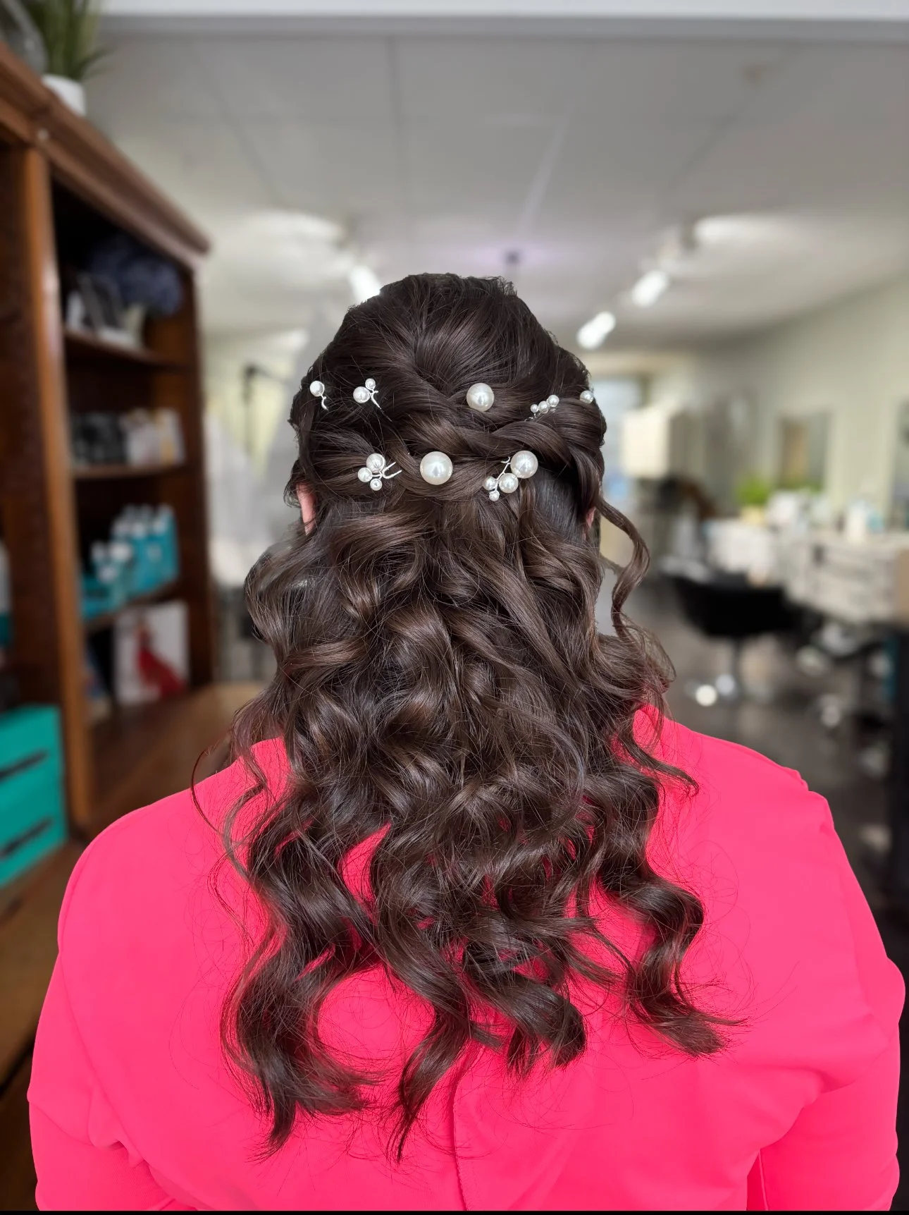 Back view of a woman with long, curly brown hair styled with pearl and silver hairpins, wearing a pink top, in a room with shelves and a mirror.