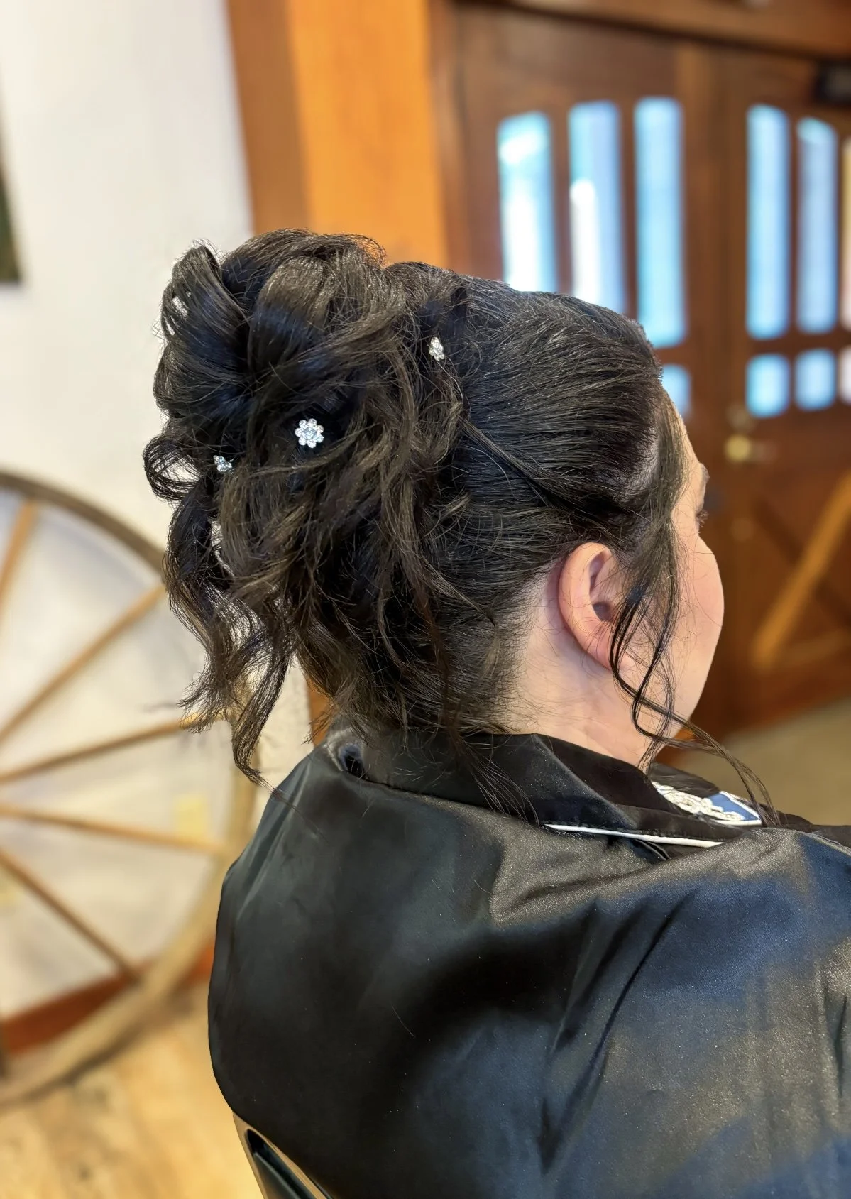 A woman with dark, curly hair styled in an elegant updo with small flower hairpins, sitting indoors near a wooden door and a decorative wheel.