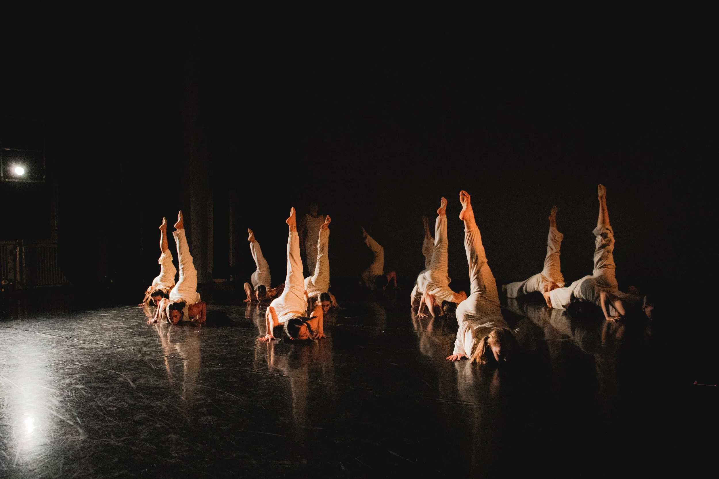 A group of nine dancers performing a synchronized dance move on a dark stage, all dressed in white outfits, with some walking on their hands and one standing in the background.
