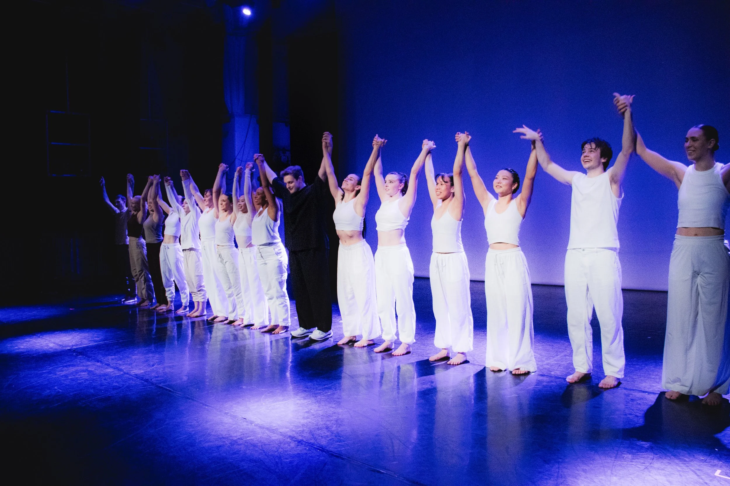 Group of performers holding hands and bowing on stage after a performance, illuminated by stage lights.