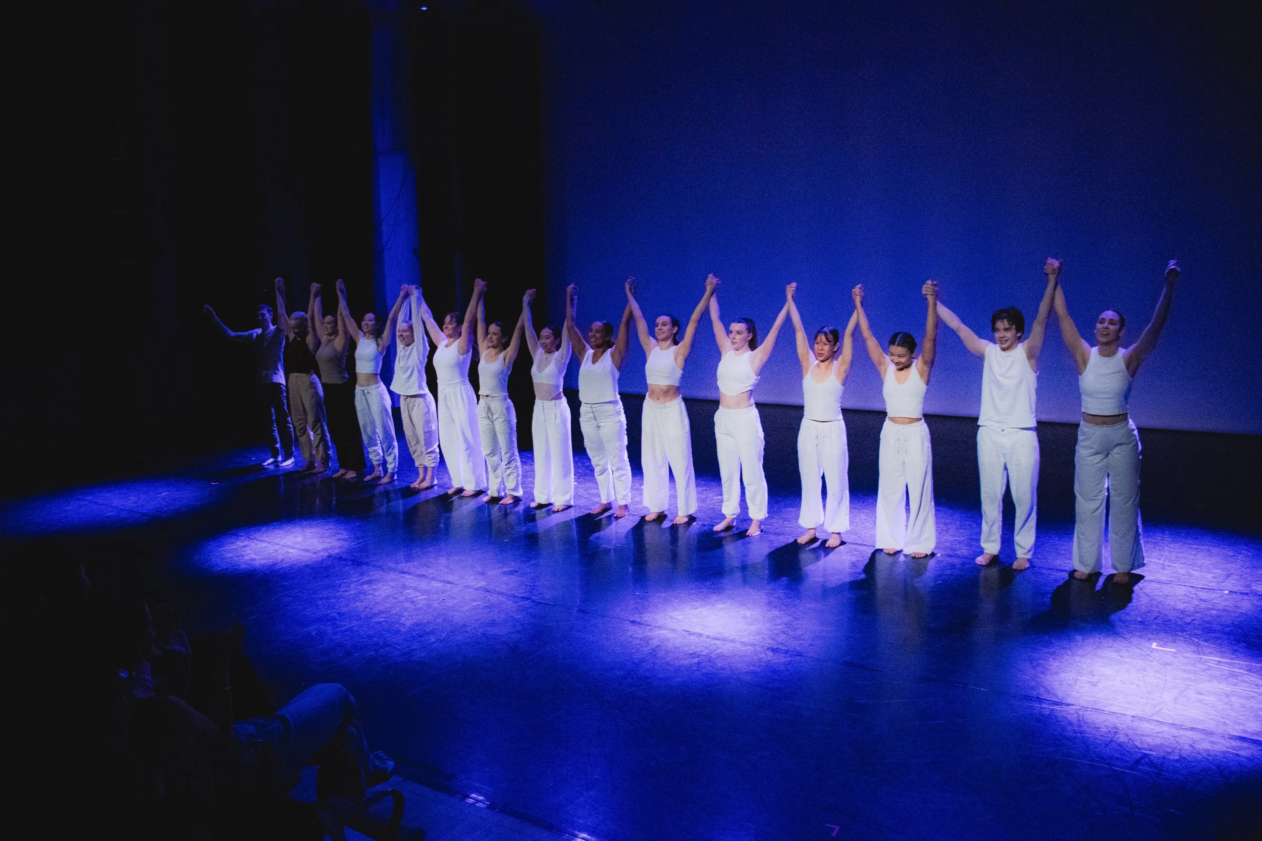 A group of performers holding hands and taking a bow on stage after a performance, standing in front of a plain blue background, dressed in white and dark costumes.