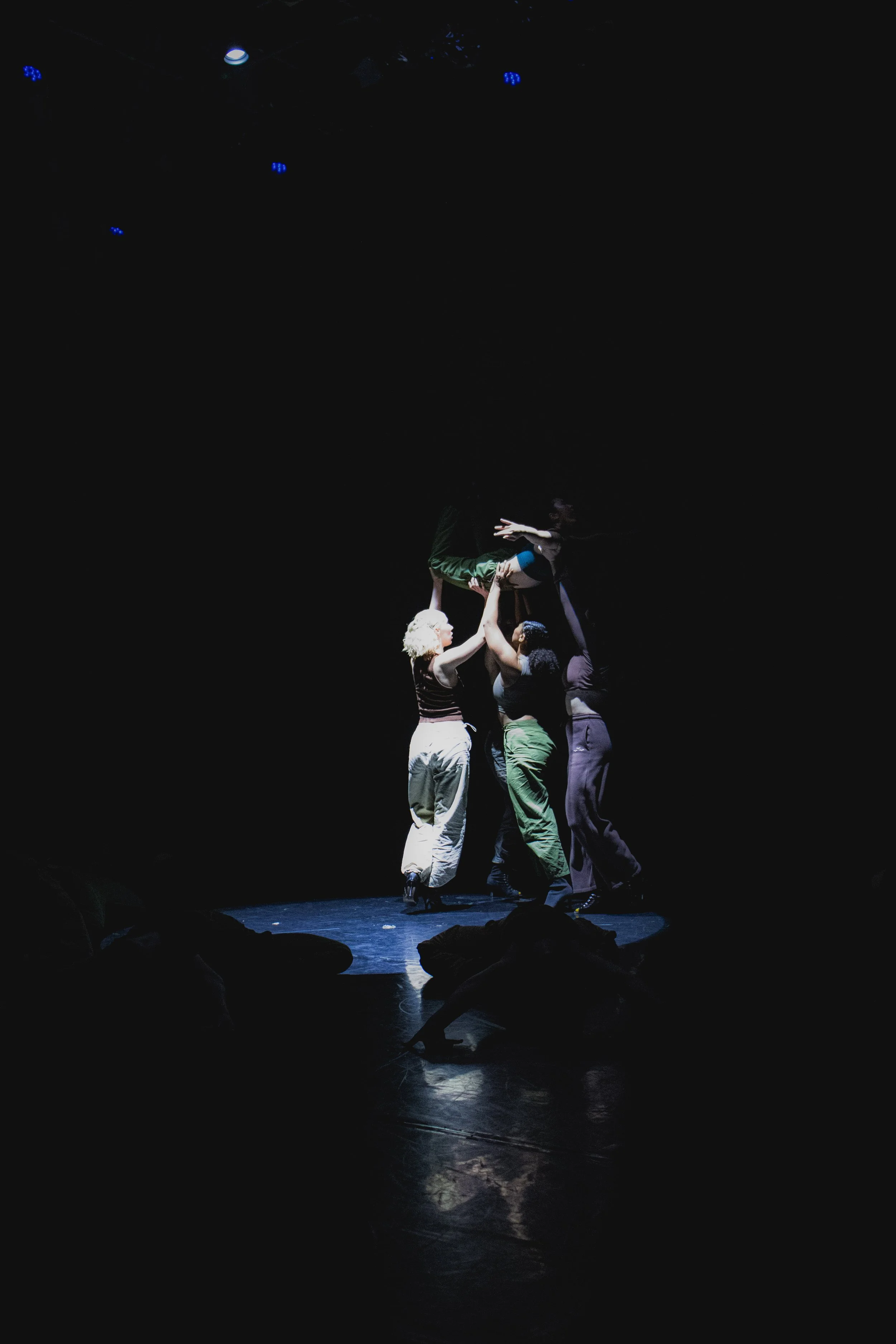 Performers on stage, three standing and lifting one person above their heads in a dark setting with spotlight, stage lights visible.