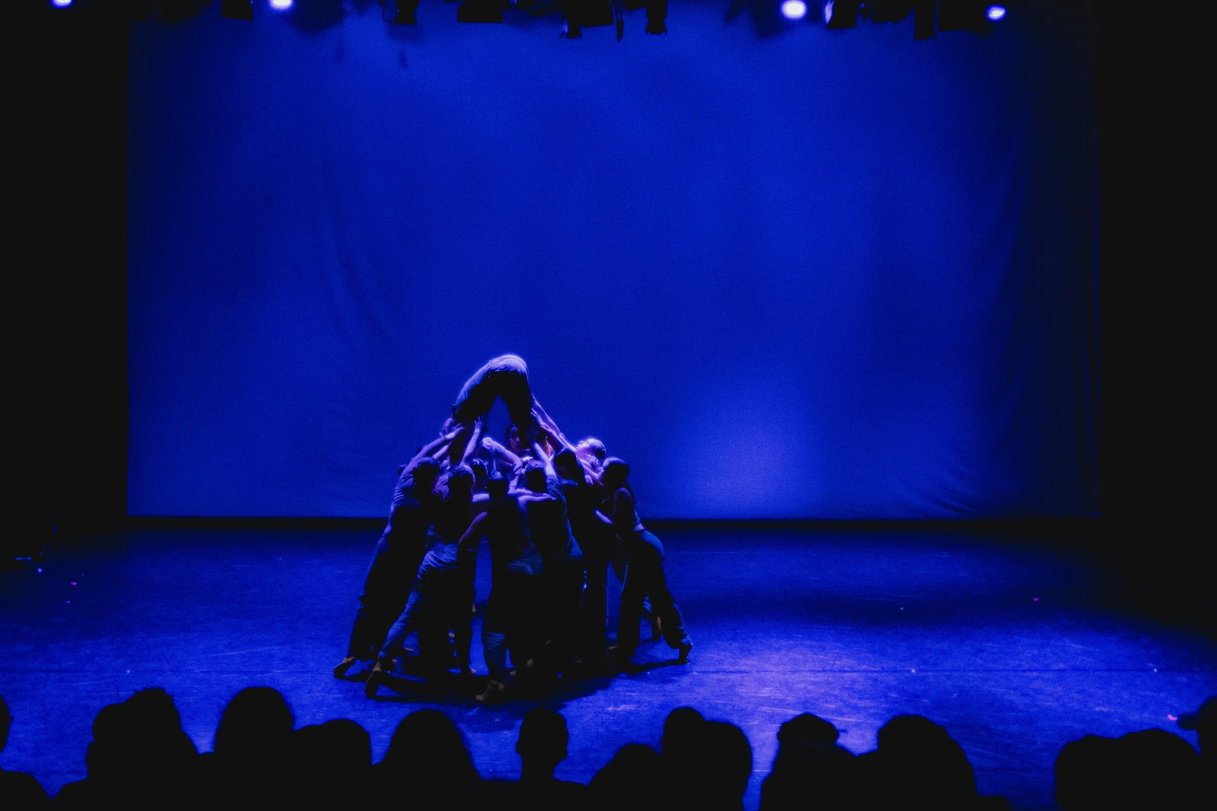 Group of performers on stage under blue lighting forming a pyramid shape with one performer at the top, in front of a plain blue backdrop, with audience silhouettes in the foreground.