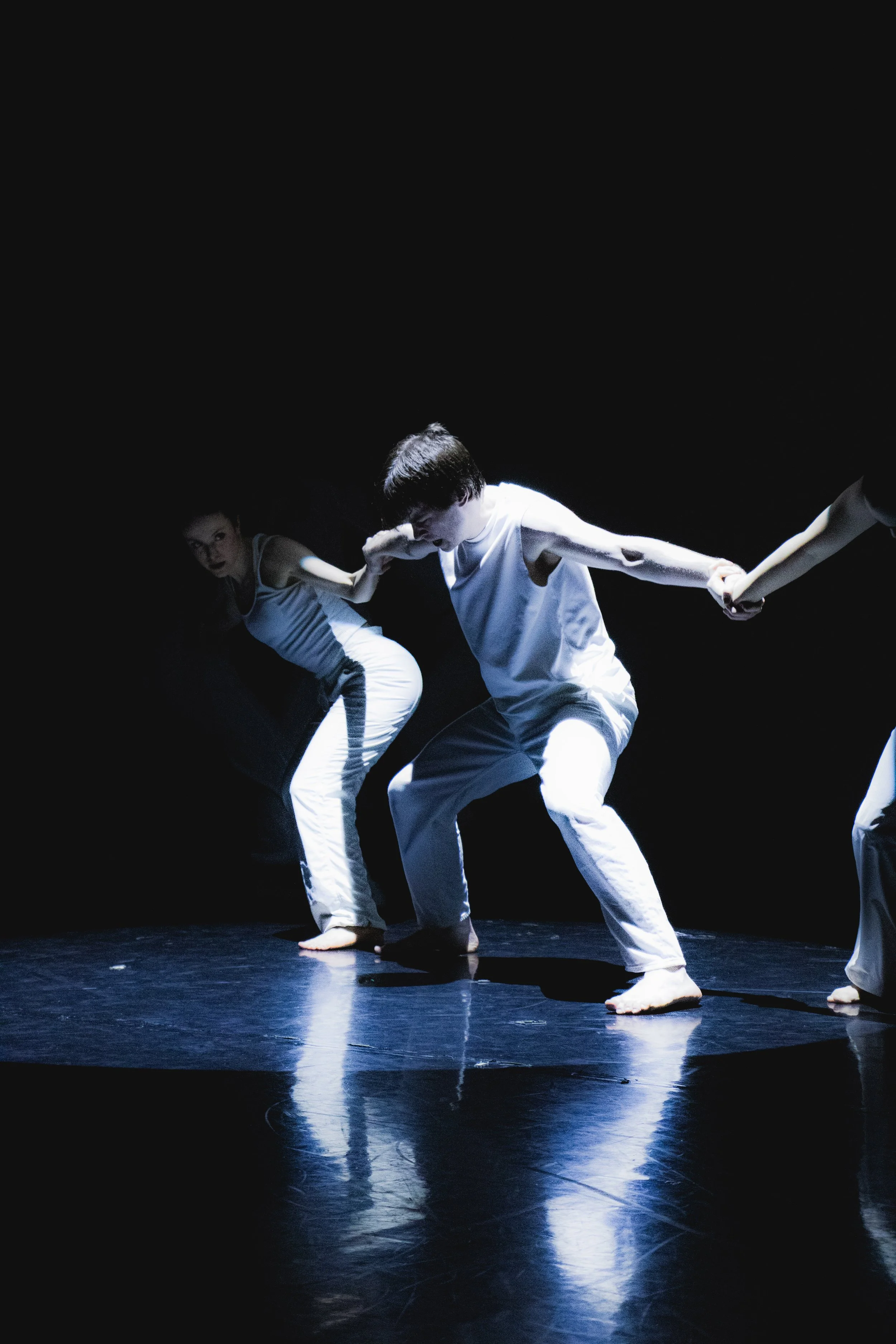 Dancers in white costumes perform on a dark stage, holding hands.