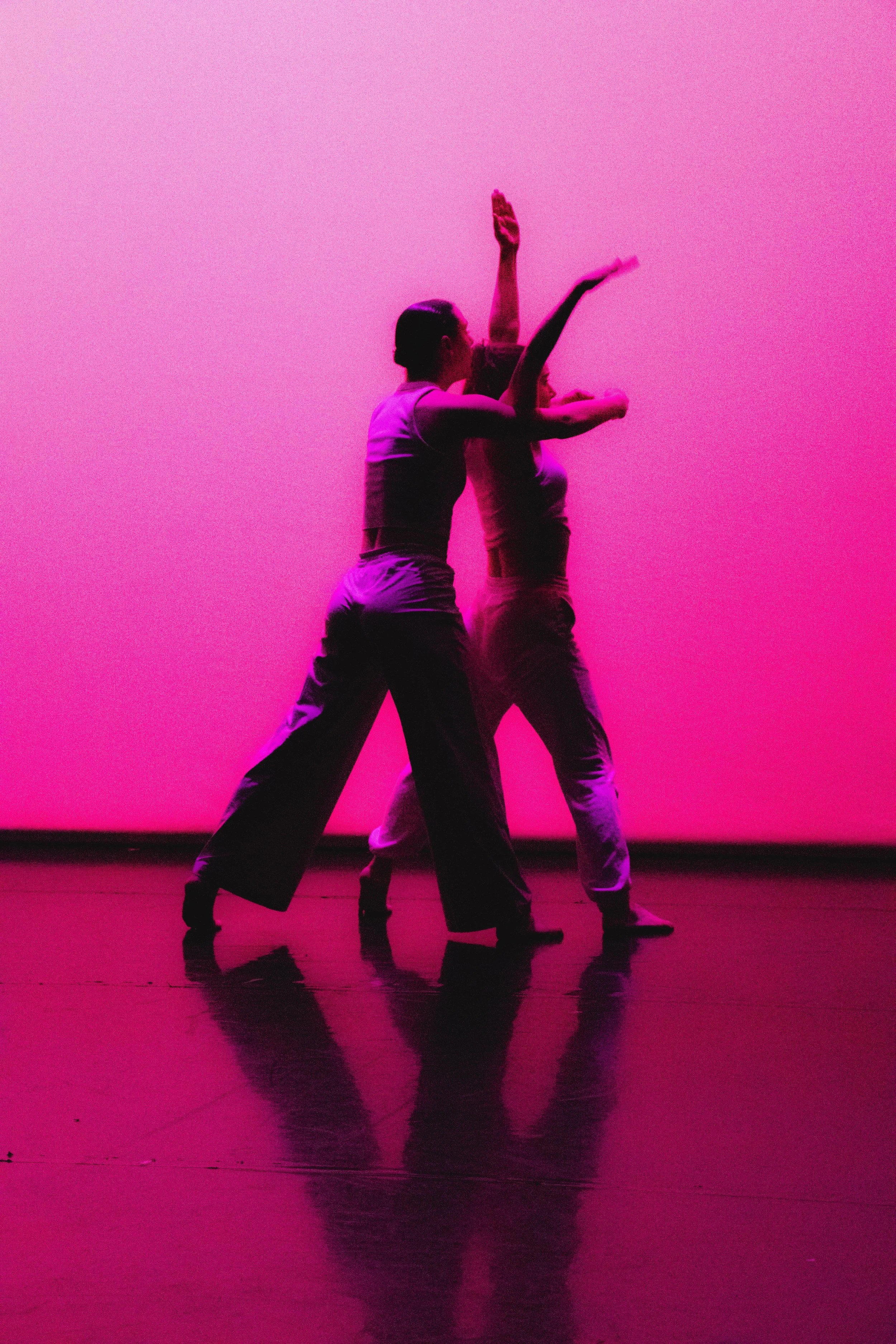 Two dancers perform on stage with a pink background. One dancer is leaning back while the other supports them, both with arms extended. Their reflections are visible on the glossy floor.
