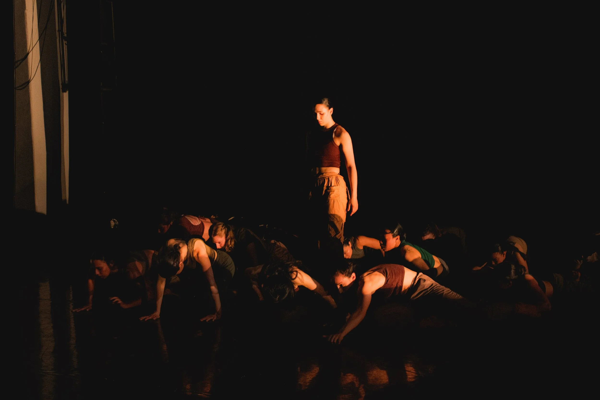 A young woman standing on stage above a group of people bowing in an act of performance or acknowledgment, with a dark background.