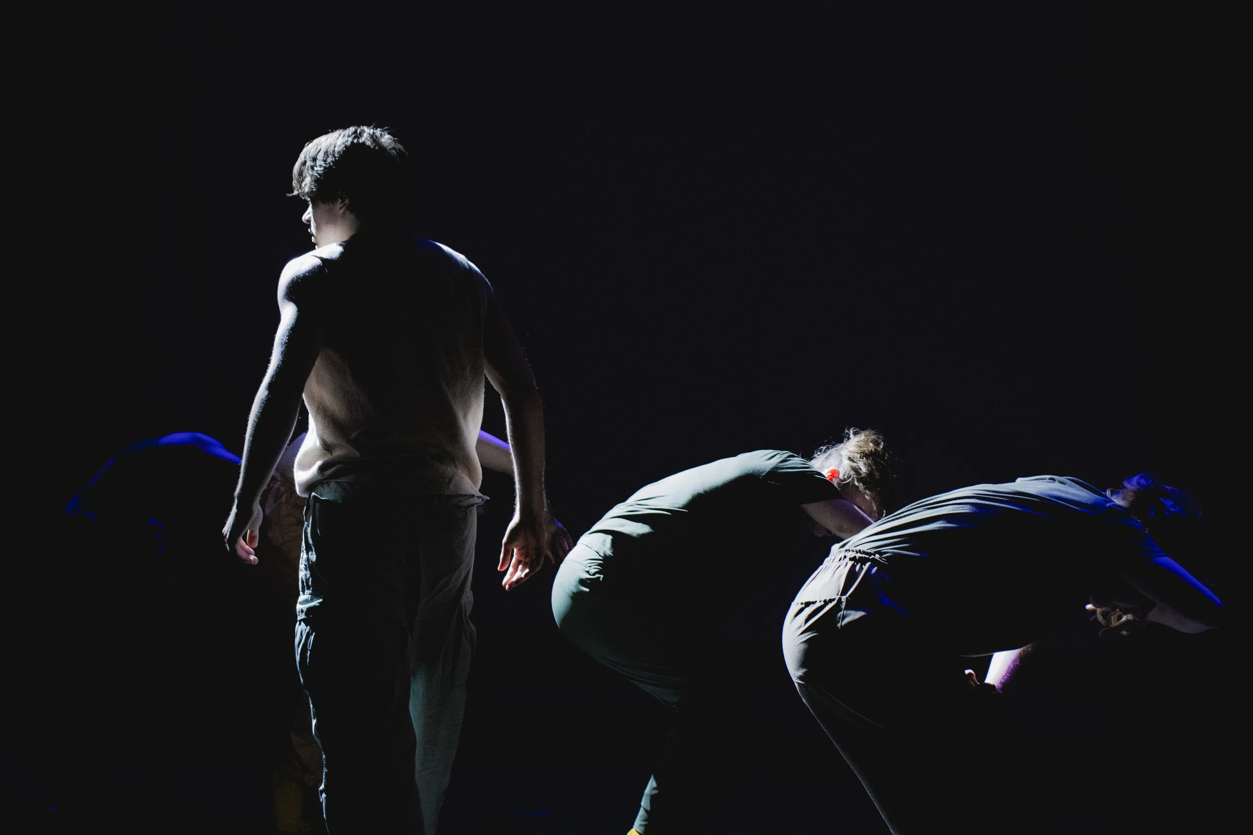 Dark stage with four people, one standing and three bending over, illuminated by spotlights, in an artistic performance or rehearsal.