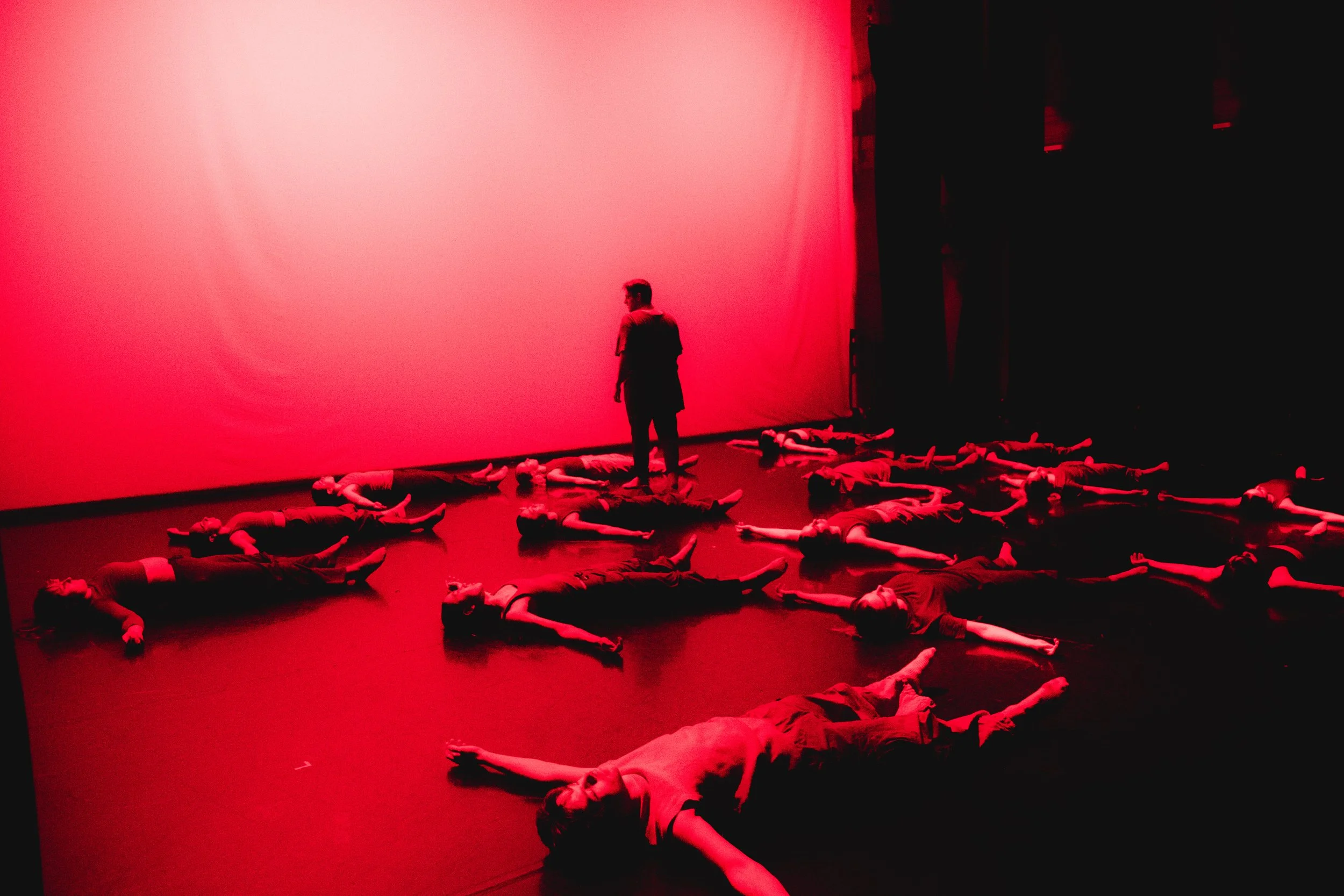 A person standing in a dark room illuminated by red light, surrounded by multiple people lying on the floor in various poses.