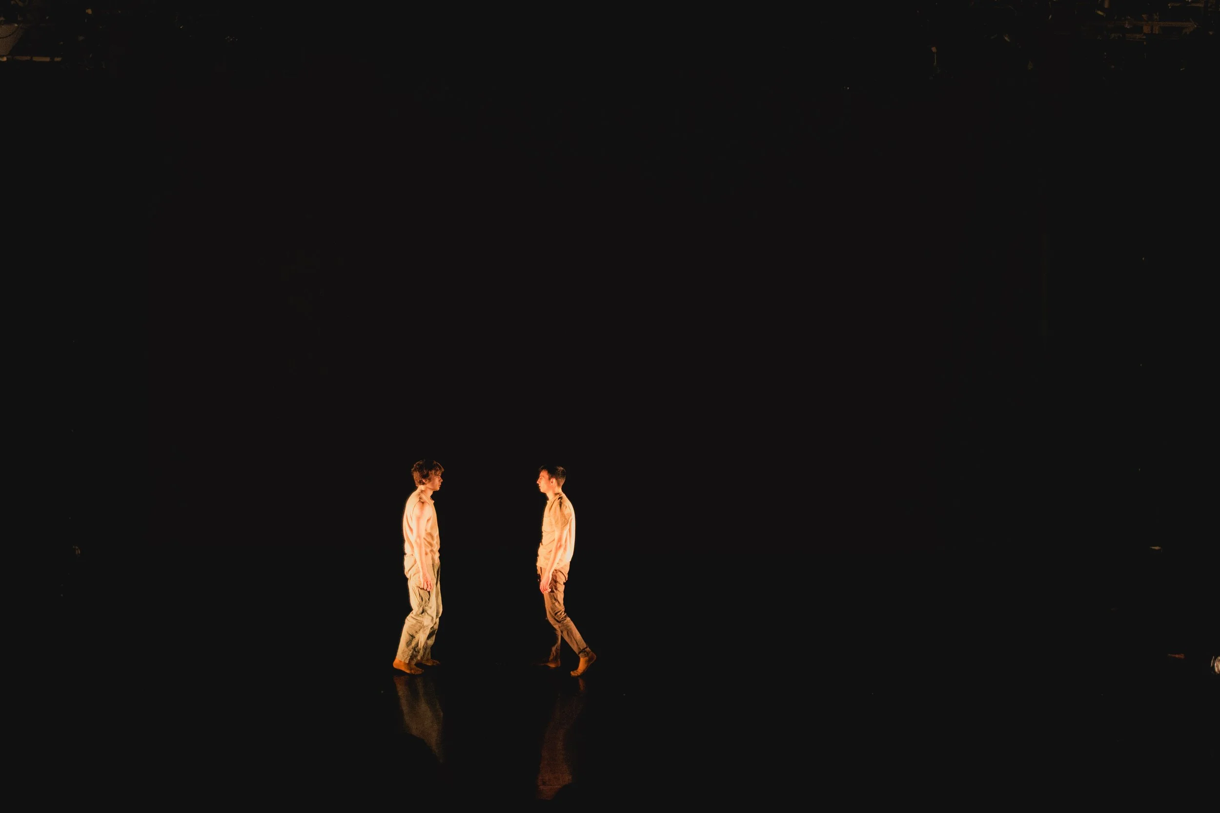 Two people standing on a dark stage facing each other, illuminated by warm stage lights, with a black background.