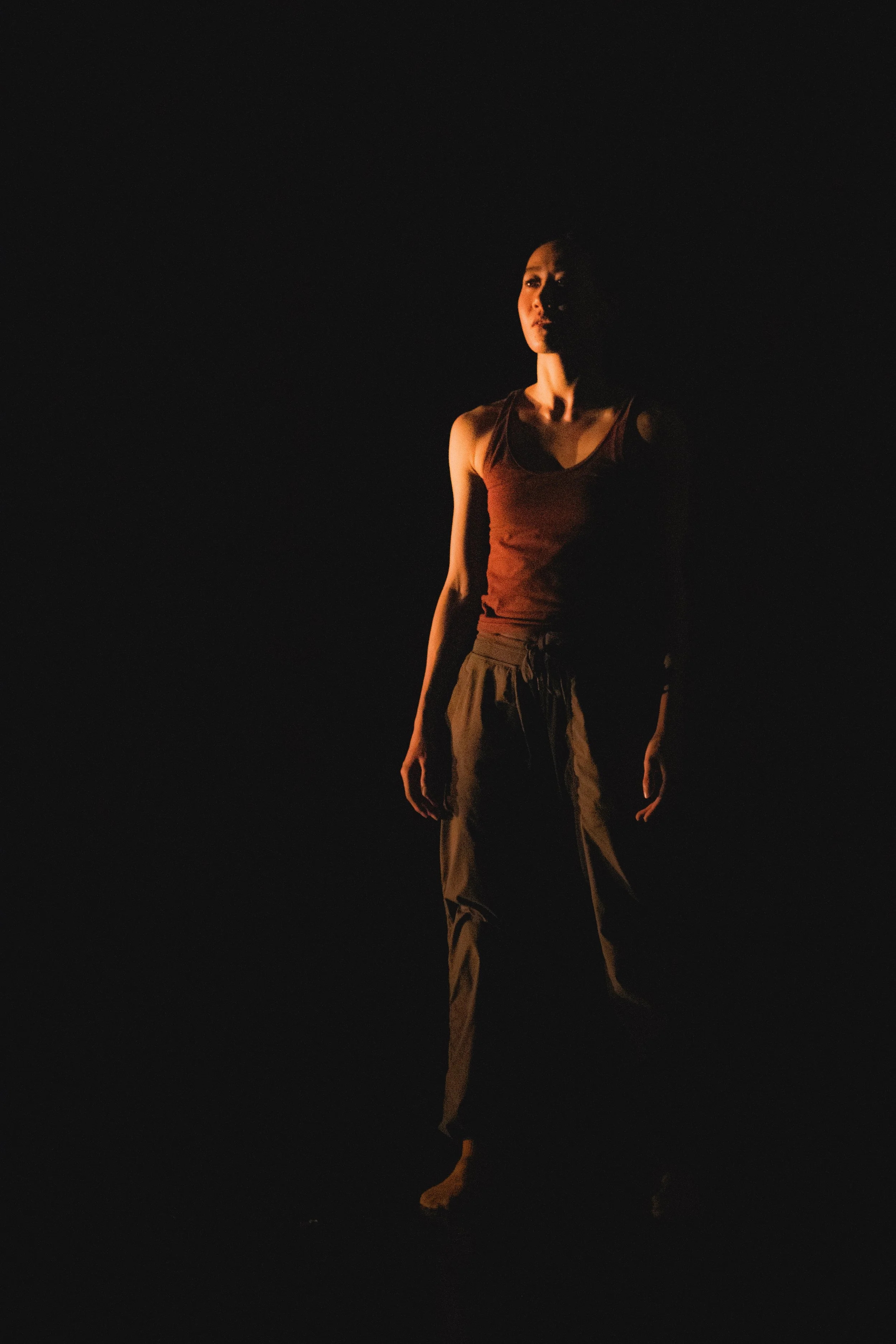 A woman standing in a dark space with minimal lighting highlighting her face and upper body, wearing a sleeveless top and loose pants.