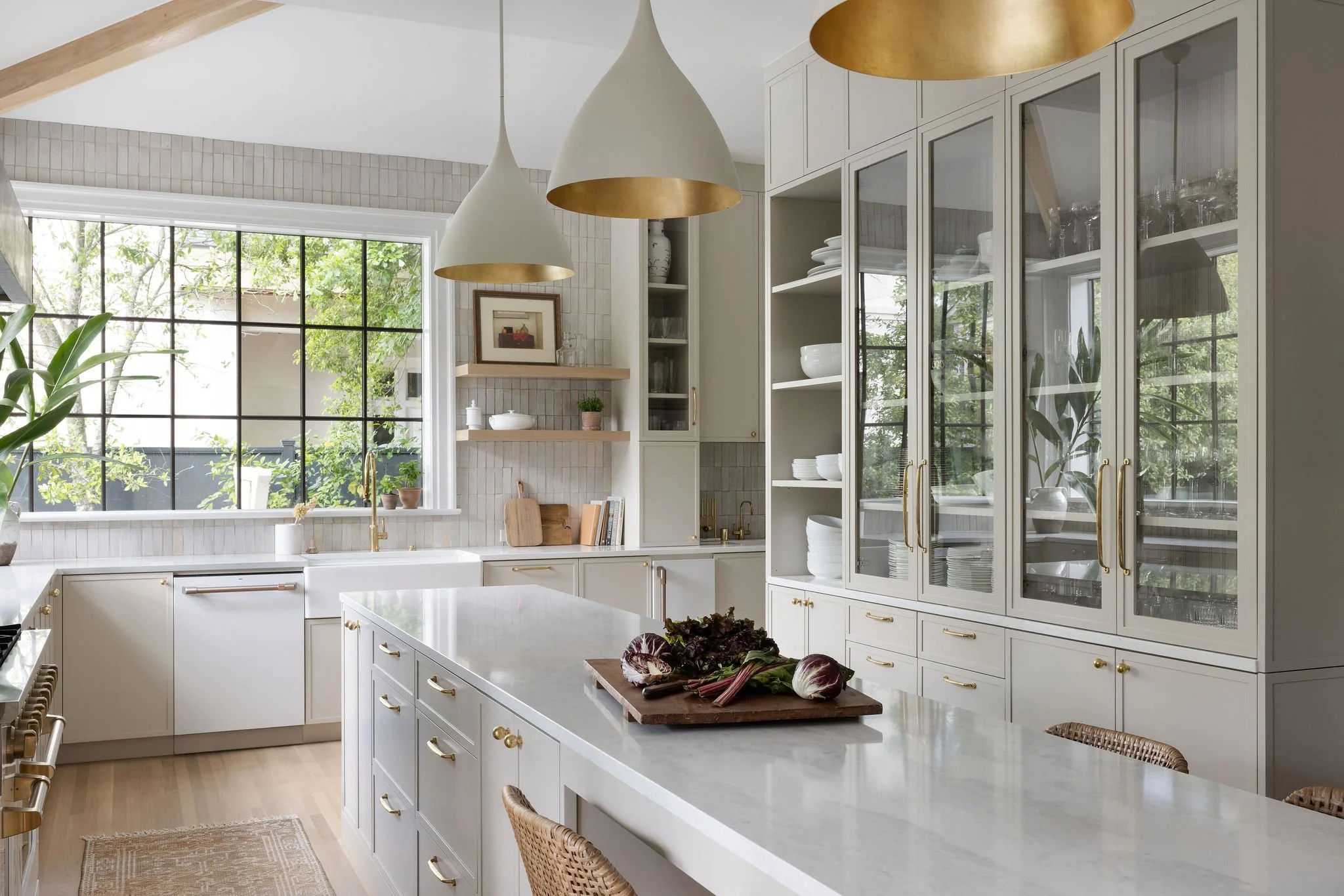 Modern kitchen with white cabinets, marble countertops, large window with black gridpanes, gold hardware, pendant lights, and fresh vegetables on the island.
