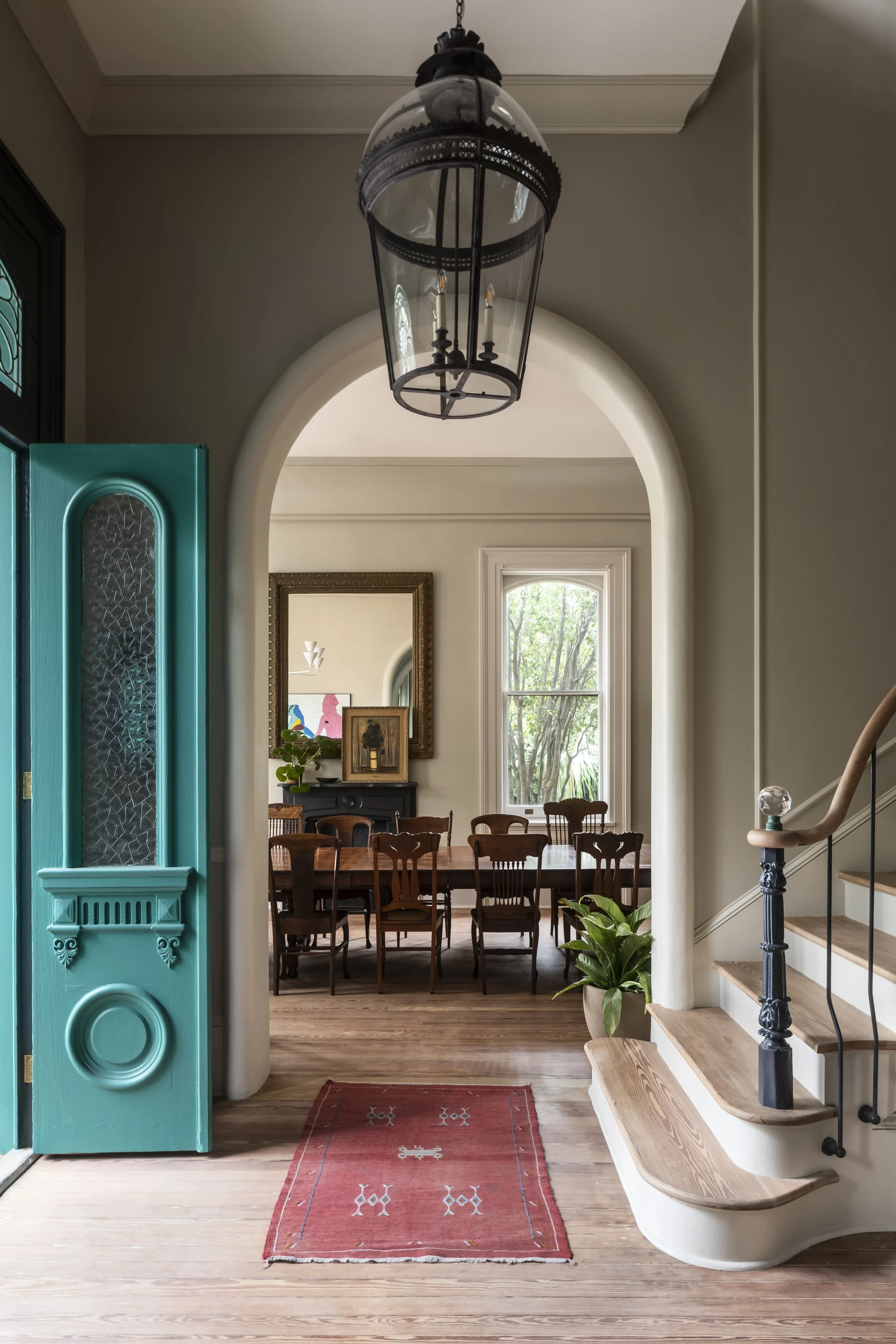 View of a warm, inviting dining area in a house, seen through an open turquoise door. The interior features a rectangular wooden dining table with wooden chairs, a large window with natural light, and decorative artwork on the walls. There is a vintage chandelier hanging from the ceiling and a red area rug on the wooden floor, alongside a staircase with wooden steps and black wrought-iron balusters.