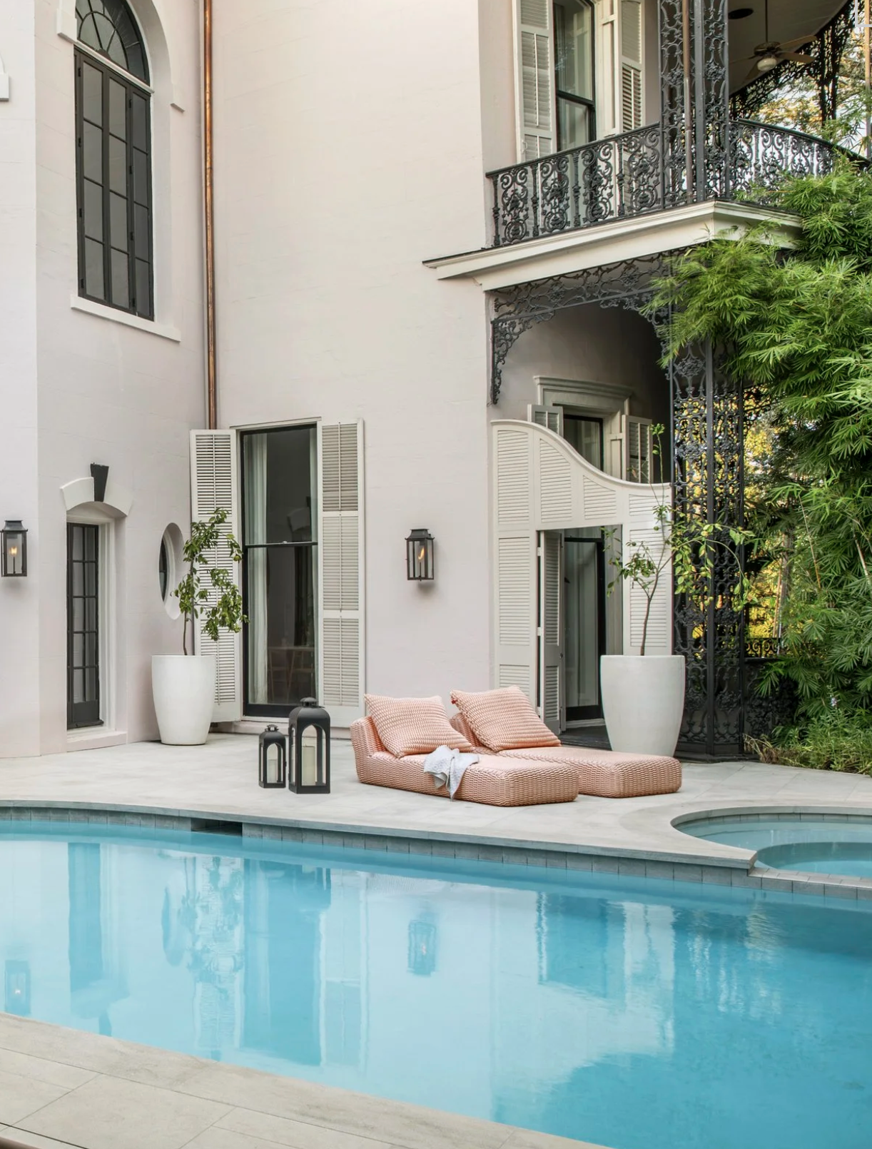 An outdoor patio of a house with a swimming pool, pink lounge chairs with pillows, large potted plants, black lanterns, and an ornate iron balcony above. The house has tall windows and white shutters.