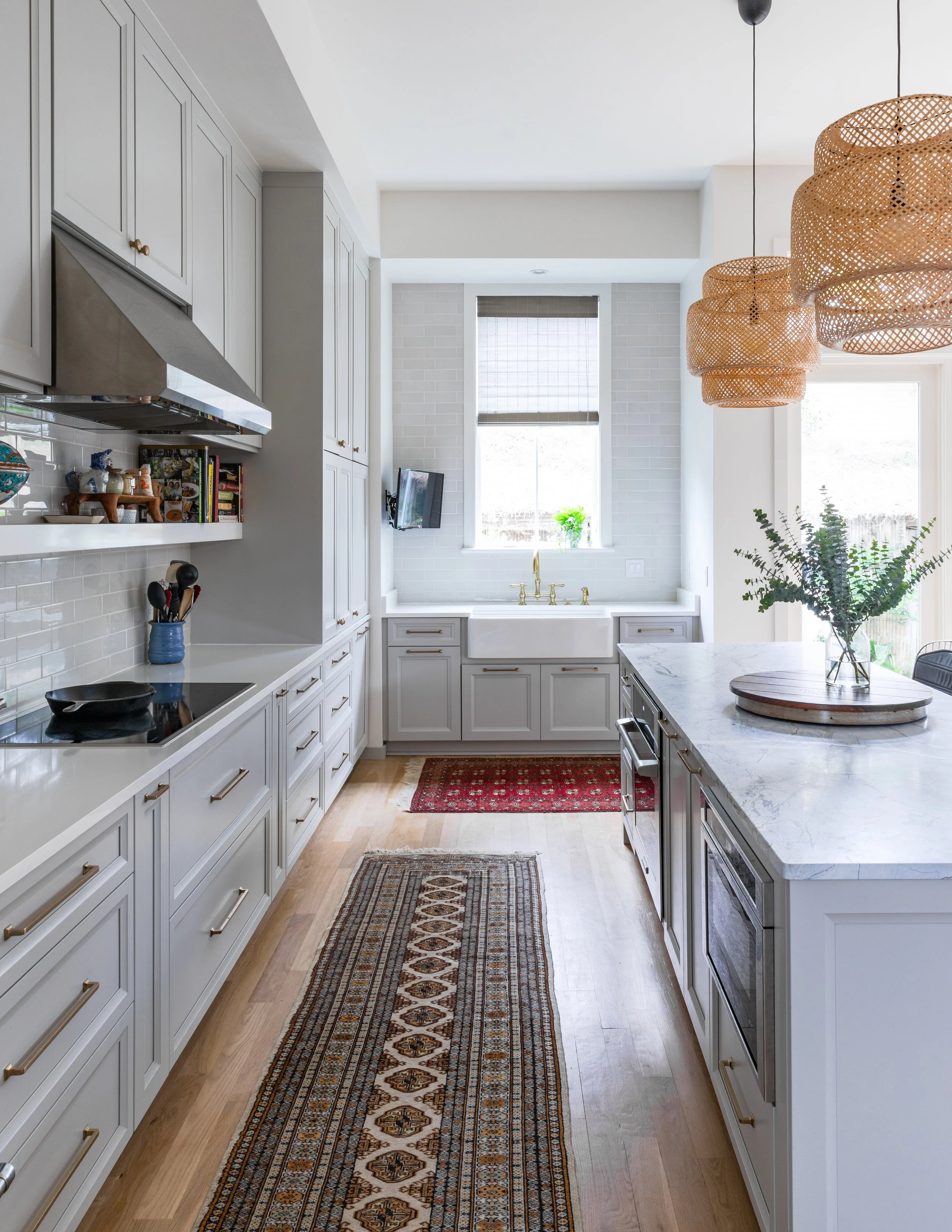 A bright, modern kitchen with white cabinets, a marble-topped island, a window above a white brick wall, hanging woven pendant lights, a small television on the far wall, and a floral dish on the counter.