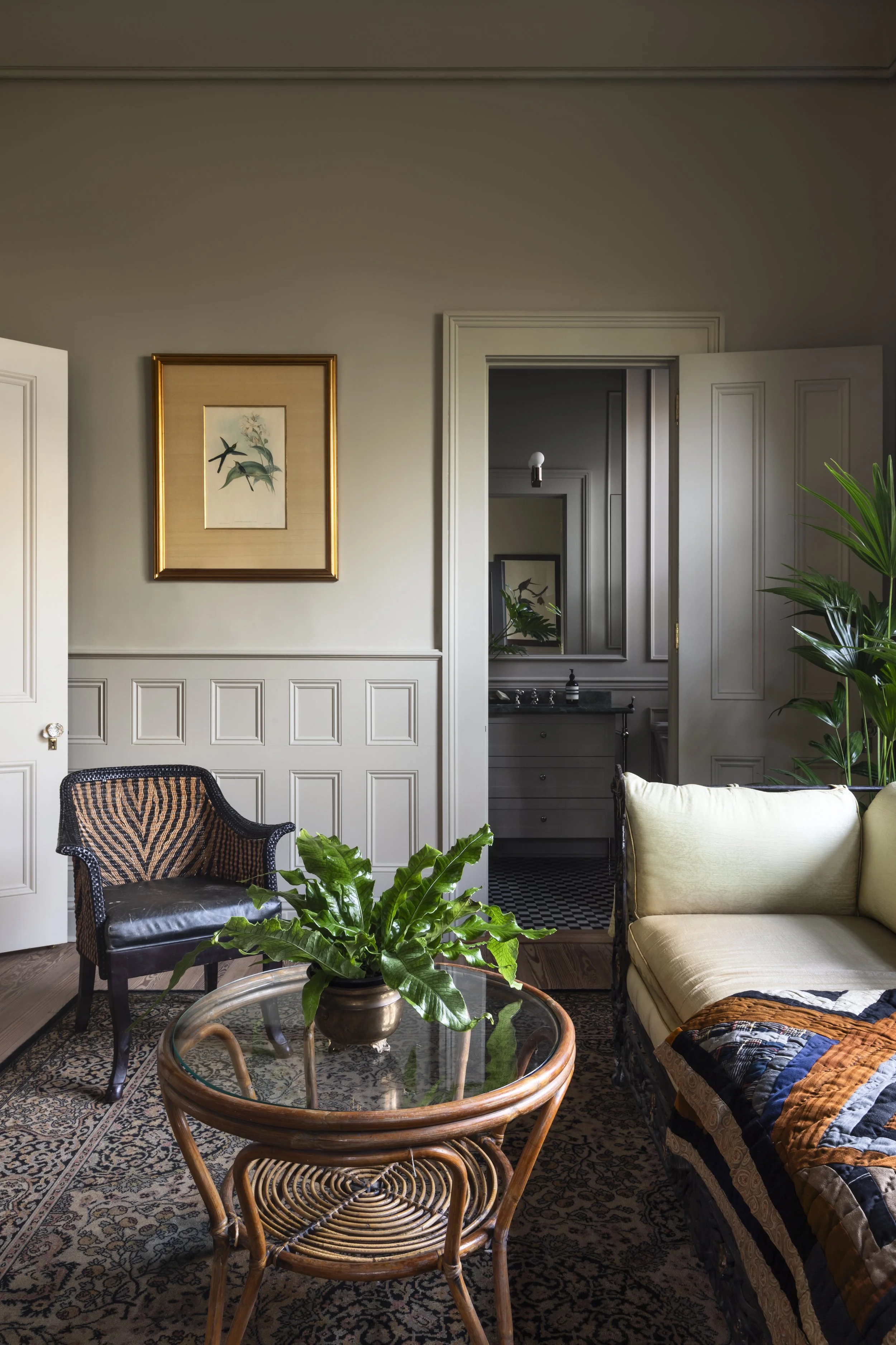 A cozy living room with a yellow sofa, a wicker chair, a glass-top coffee table with a potted plant, framed artwork on the wall, and a mirror reflecting another room with a small sink and plants.