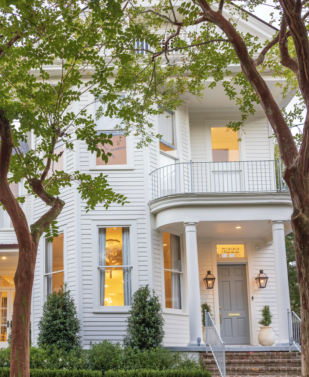 A white, multi-story house with a rounded porch supported by columns, front steps, and a gray door. Windows glow with warm yellow light, and there are two large potted plants near the entrance. The house number 5222 is visible above the door. Two tre