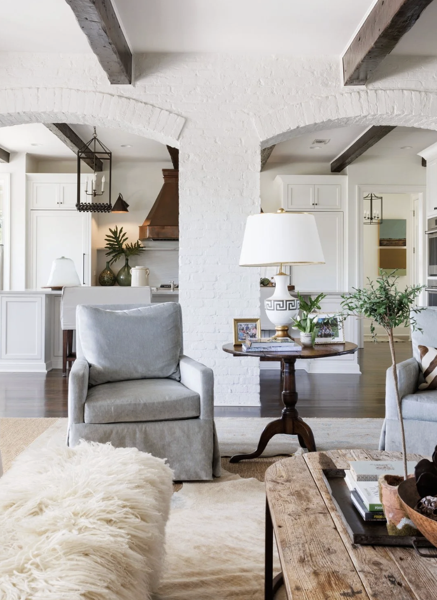 Living room with white walls, a gray armchair, a wooden side table with a white lamp, and a wooden coffee table with decorative items. Visible kitchen area in the background with white cabinets, a copper hood, and green plants.